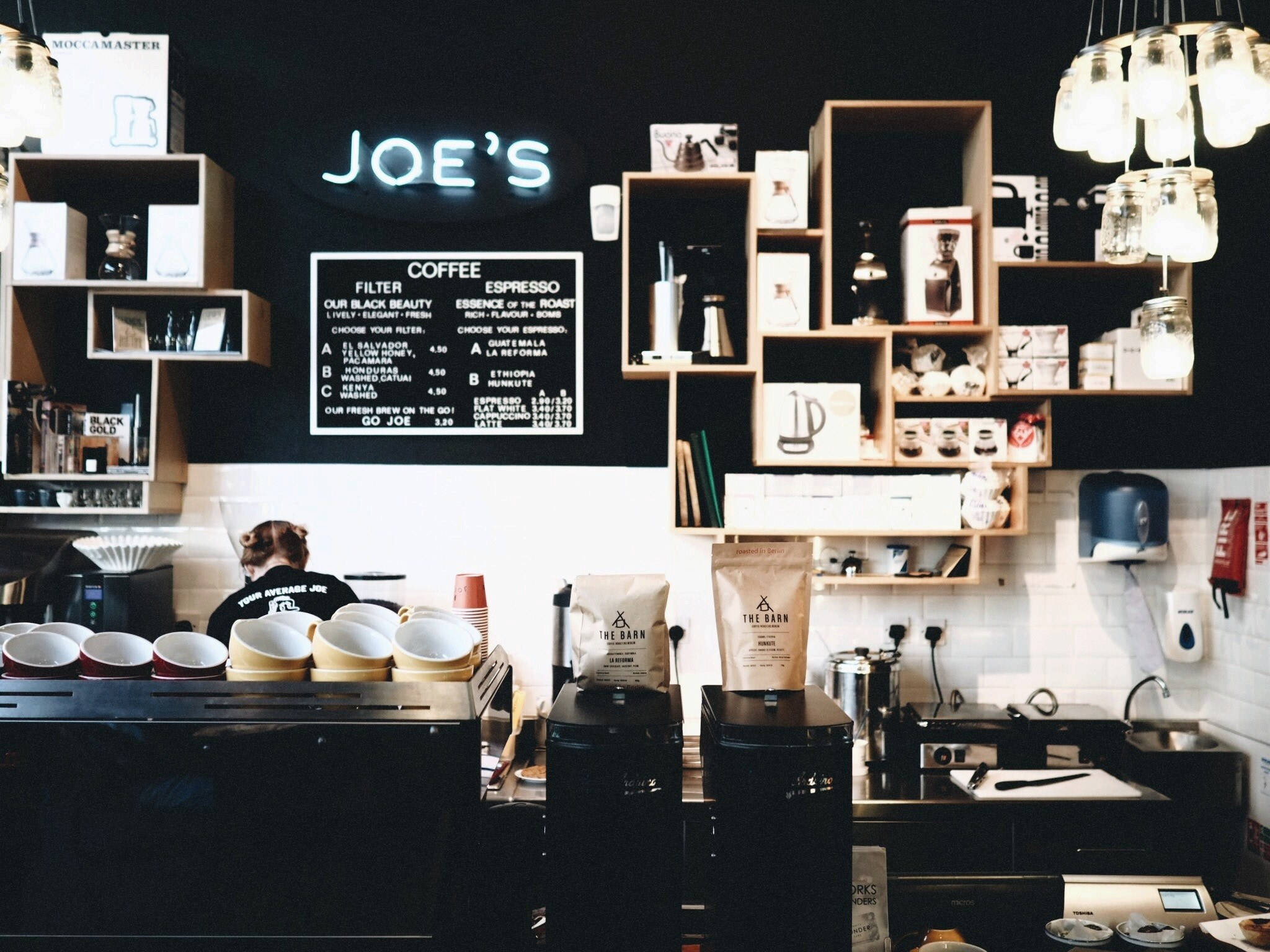 Coffee Shop 桌布 壁紙 — 舒適的咖啡店內部，吧檯後有一位咖啡師，上方是發光的霓虹燈牌，寫著 JOE'S。
