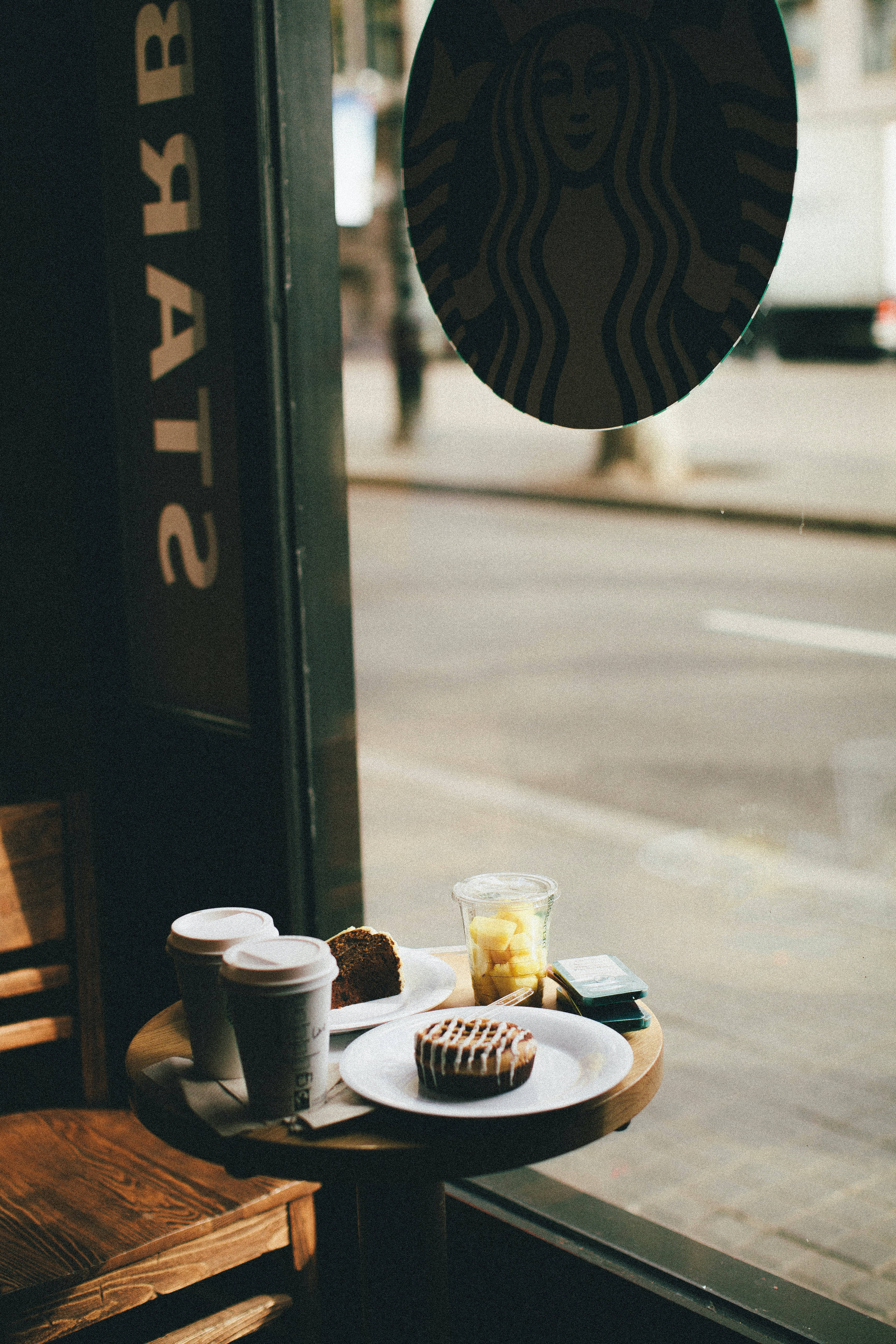咖啡飲品和糕點放在咖啡店窗邊的木桌上，窗外有Starbucks標誌。 — 星巴克 GIF 壁紙
