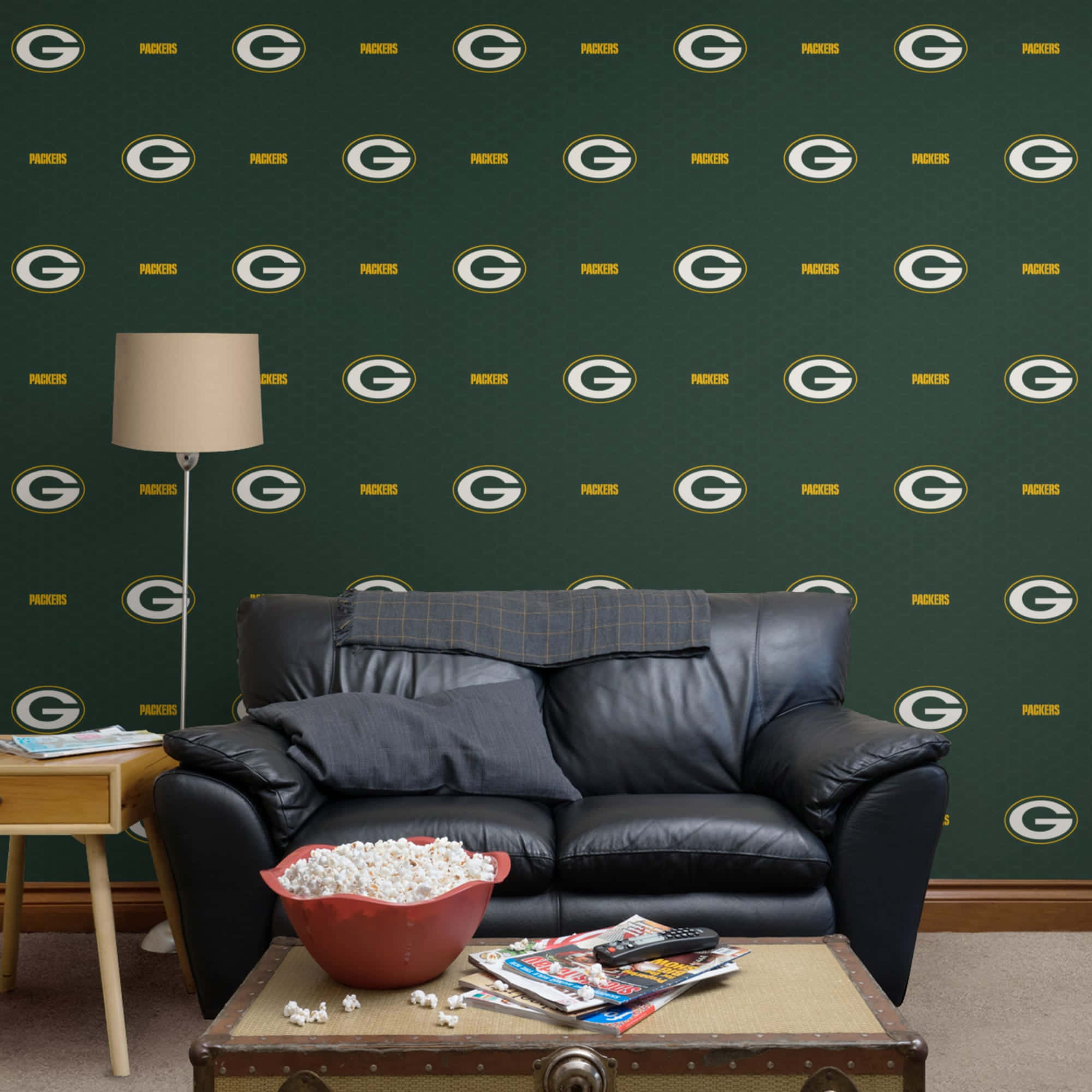 Packers 桌布 壁紙 — 舒適的客廳中，黑色皮革沙發靠著印有重複 Green Bay Packers 標誌和文字的綠色壁紙。
