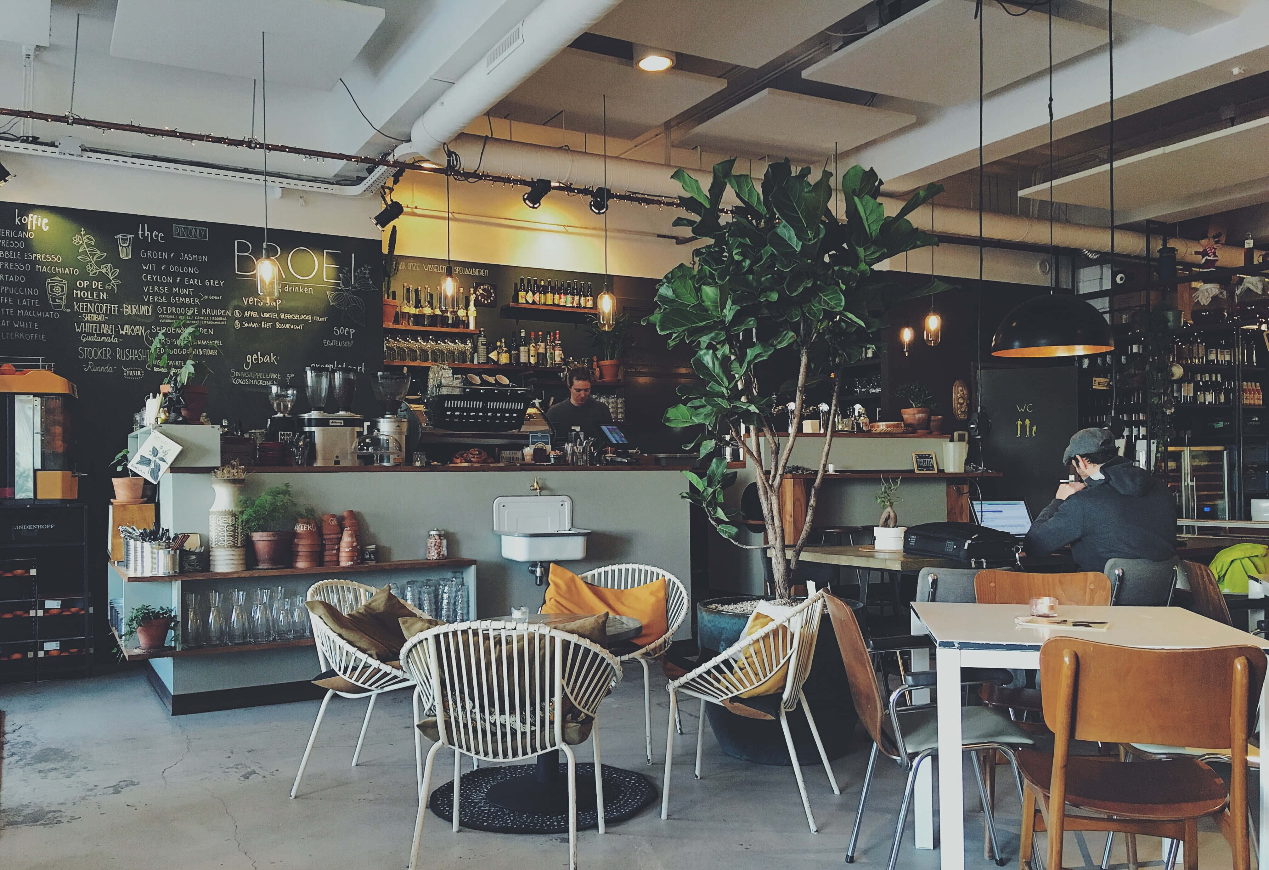Restaurant 桌布 壁紙 — 舒適的工業風咖啡館，配有外露管道、溫暖燈光、被綠色植物和黑板菜單環繞的餐桌顧客。
