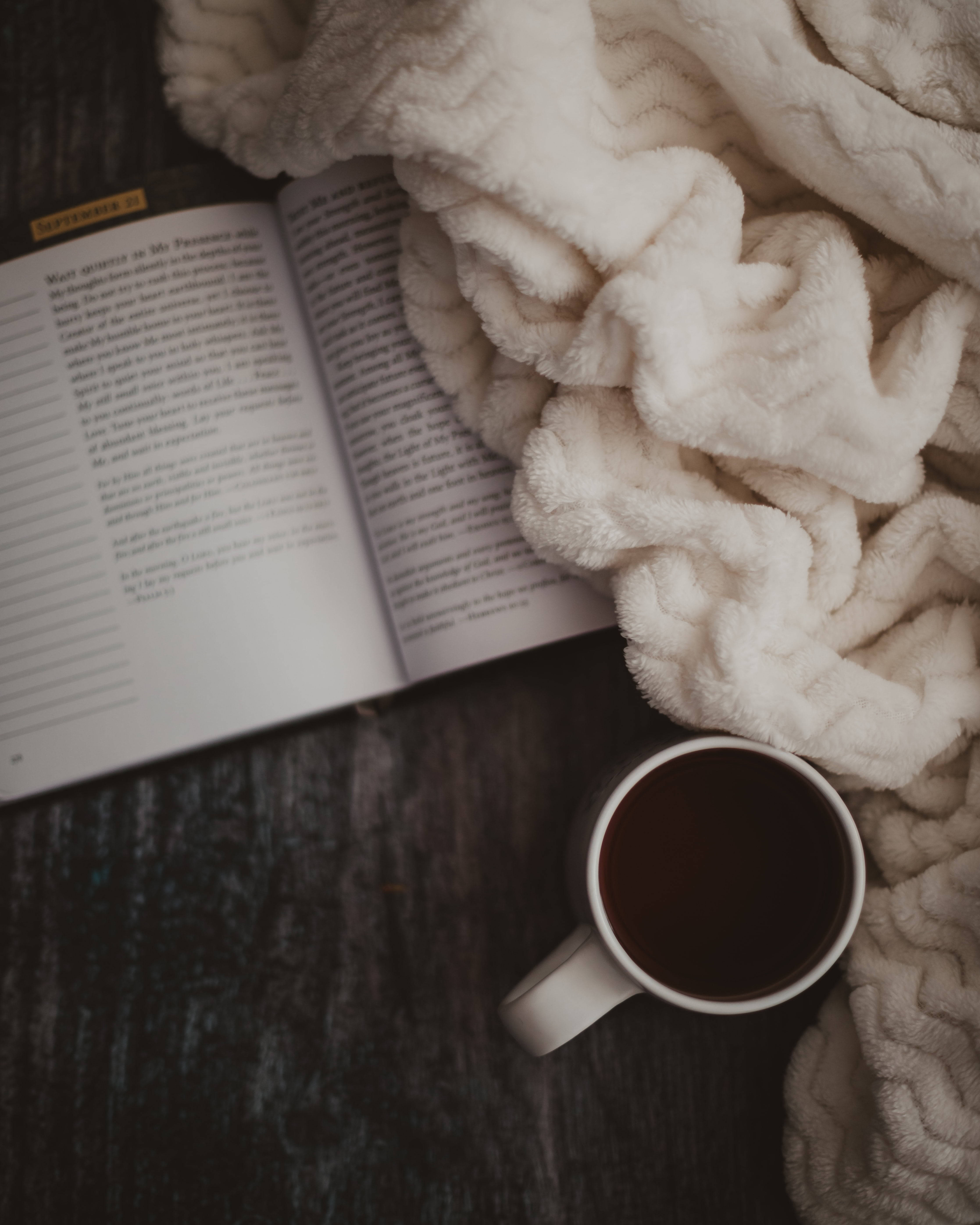 Cozy 桌布 壁紙 — 舒適的打開的書本和冒煙的咖啡杯，依偎在柔軟的白色毯子下，放在深色木質表面上。