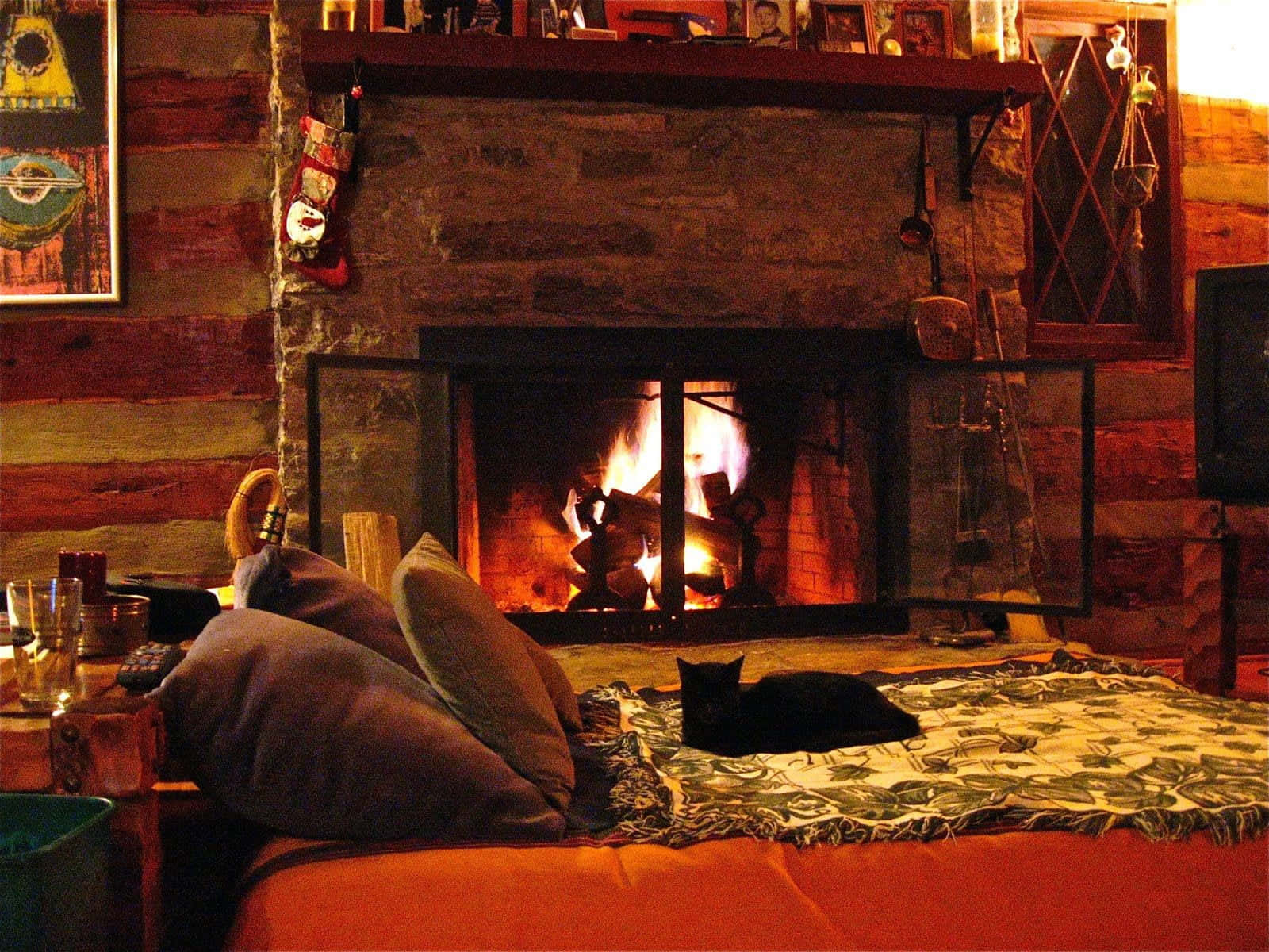 Autumn Fireplace 桌布 壁紙 — 舒適的木屋內部，設有熊熊燃燒的石壁爐，一隻黑貓躺在圖案毛毯上休息，以及溫暖的環境照明。