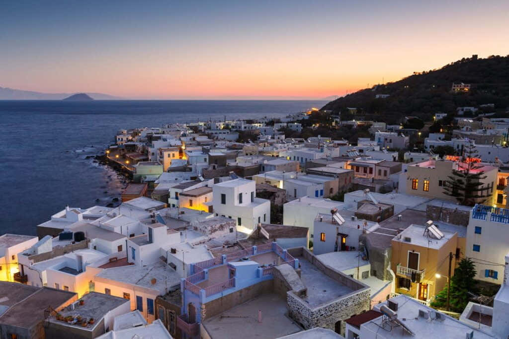 Greek Island 桌布 壁紙 — 舒適的白色村莊在日落時依偎在懸崖邊，溫暖的燈光映照著海洋地平線。