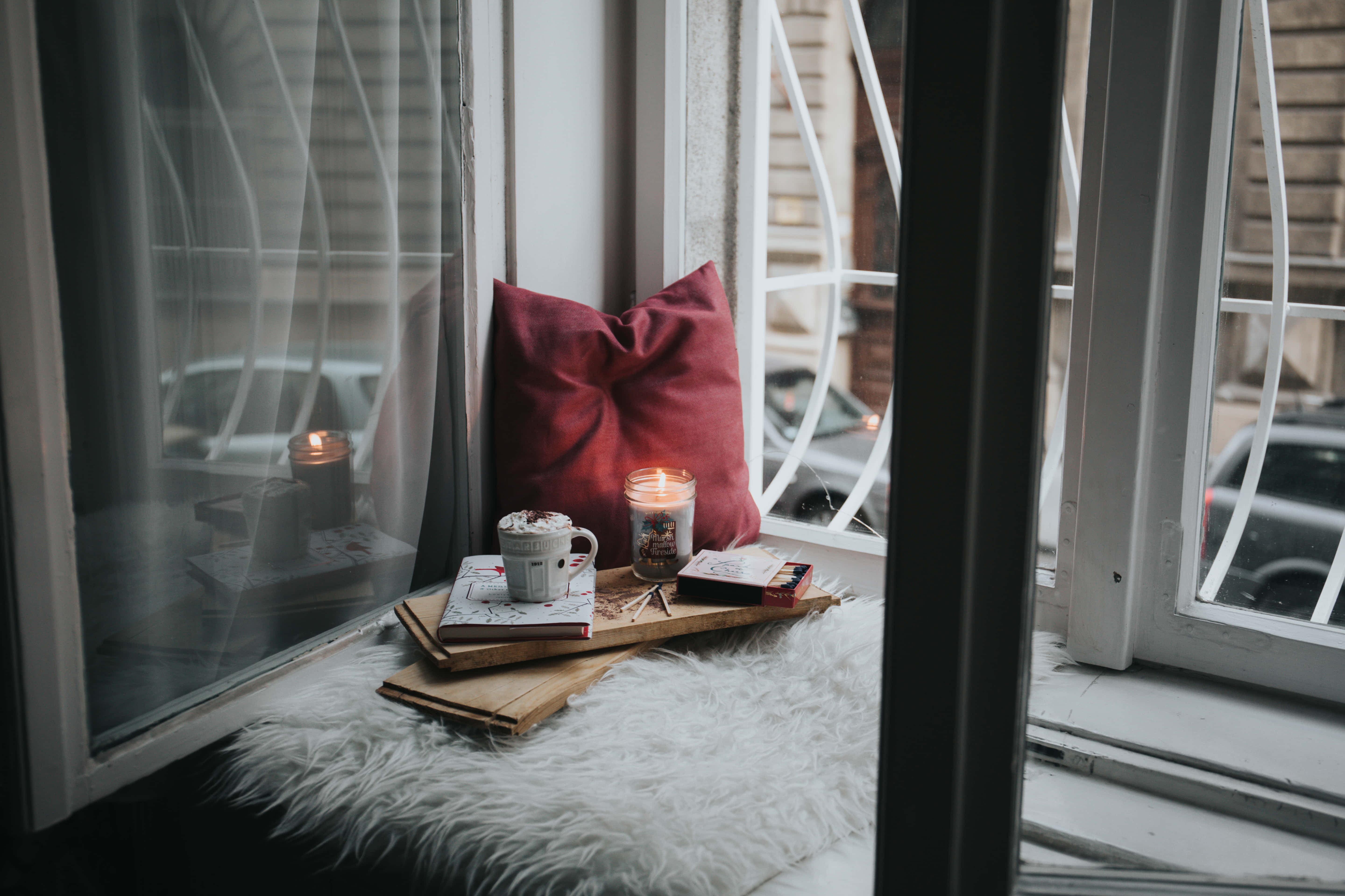 Cozy Winter Desktop 桌布 壁紙 — 舒適的窗邊座位，配有毛絨白色地毯、紅色靠墊、點燃的蠟燭和杯子，旁邊木托盤上放著打開的書本。