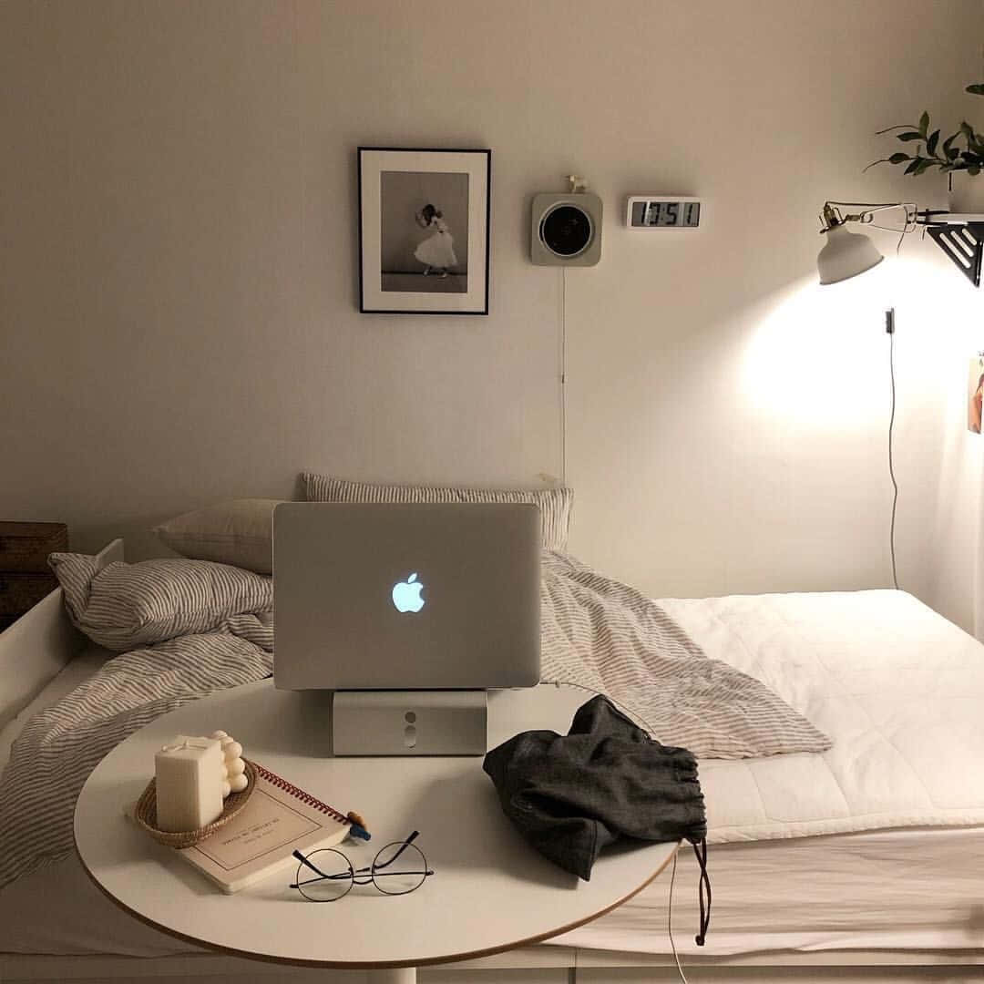 美學米色 桌布 壁紙 — 舒適的臥室角落，筆記型電腦在床邊的圓桌上發光，由溫暖的壁燈照亮。