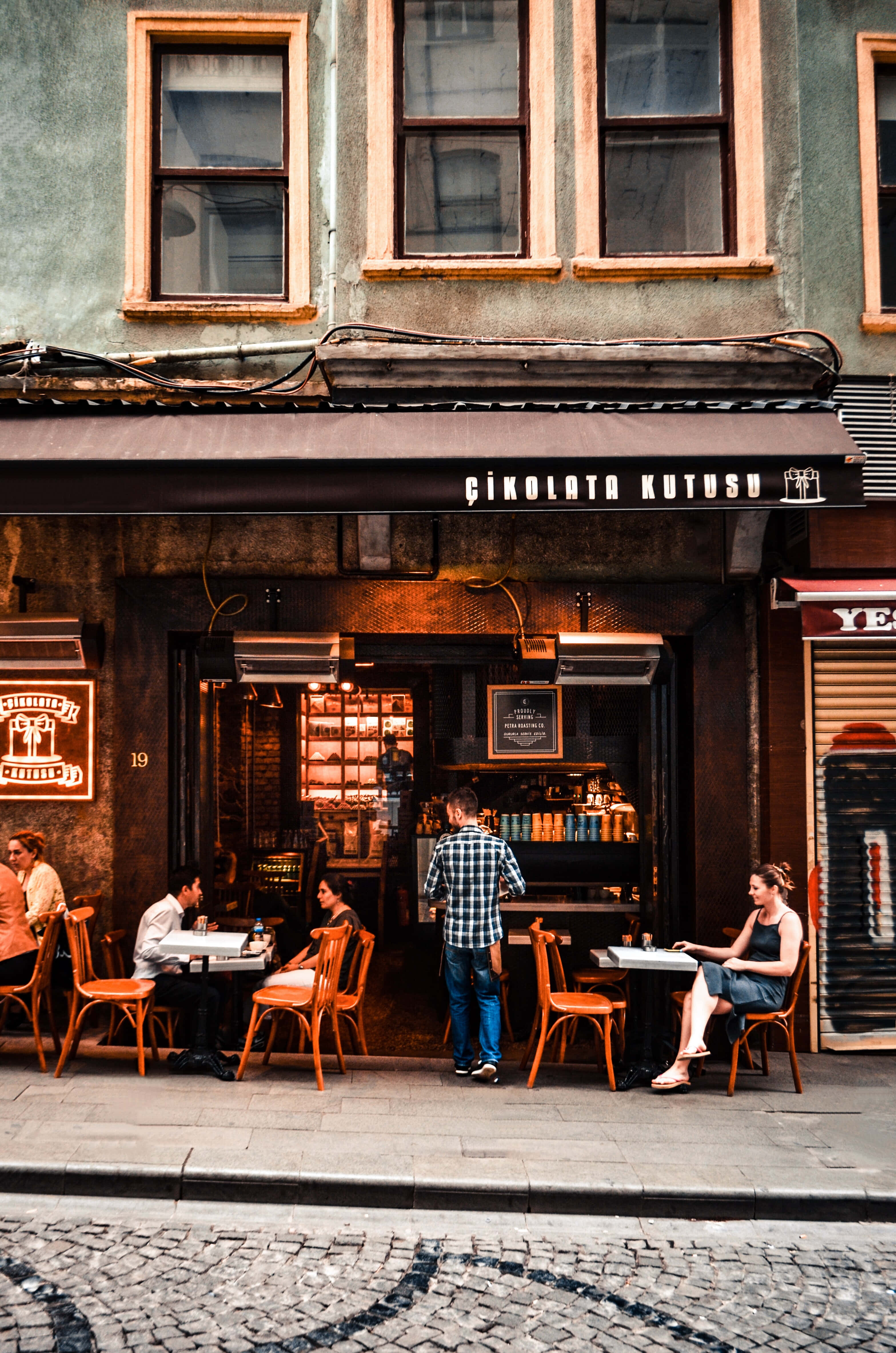 Coffee Aesthetic 桌布 壁紙 — 舒適的街頭咖啡館，顧客坐在鵝卵石人行道上的木桌旁，上方有棕色遮陽篷。