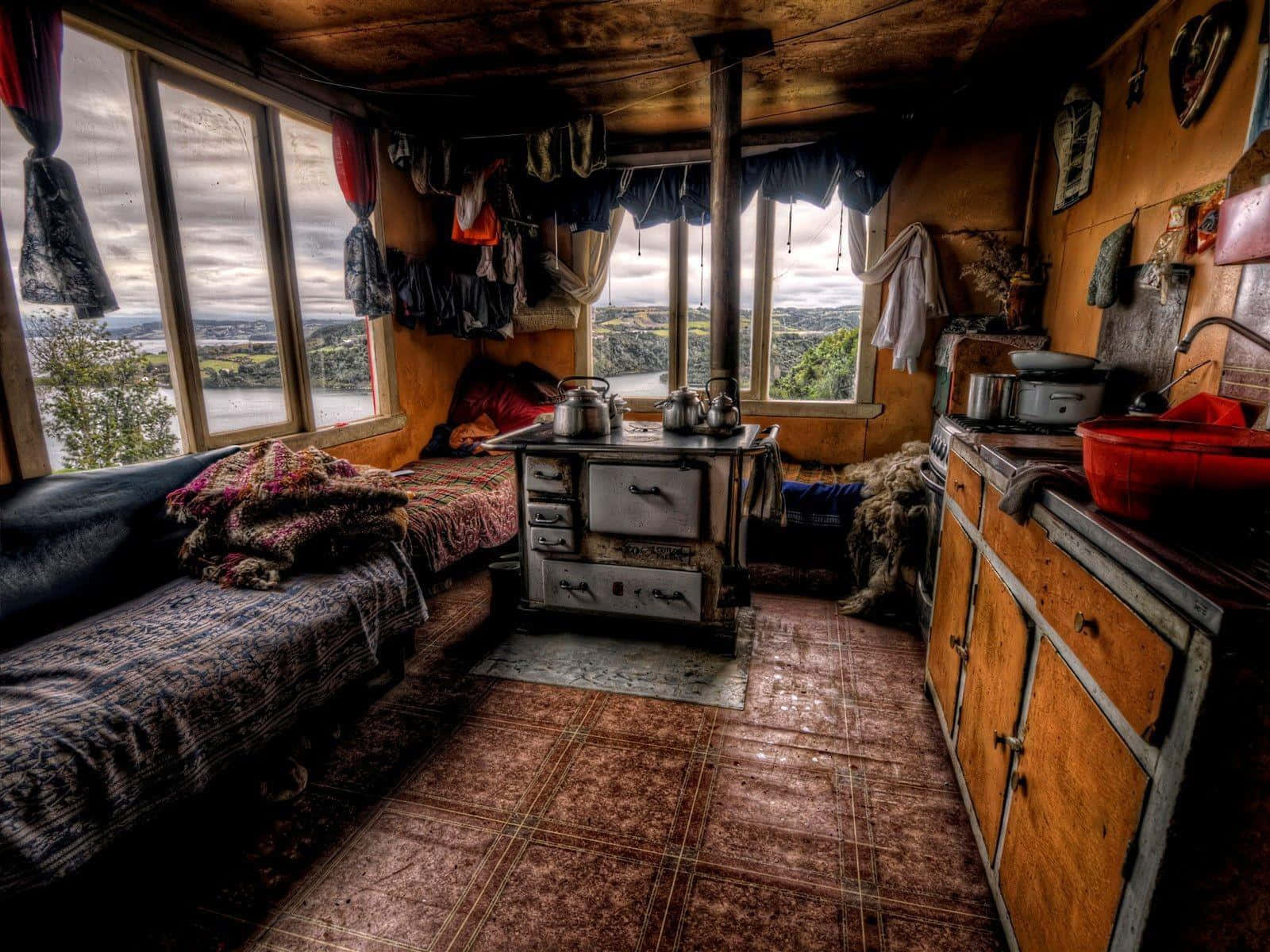 Cozy Winter Cabin 桌布 壁紙 — 舒適的鄉村小屋內部，木質牆壁和地板瓷磚，設有復古爐灶，窗戶俯瞰霧氣瀰漫的山谷，懸掛著衣物。
