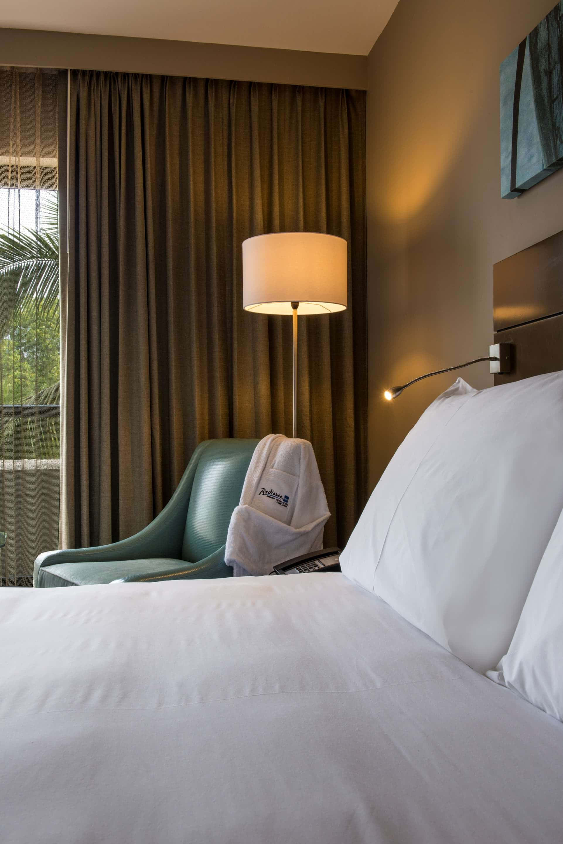 Hotel Room 桌布 壁紙 — 舒適的飯店客房，配有整齊的床鋪、掛著浴袍的藍綠色椅子，以及來自落地燈和壁燈的溫暖燈光。