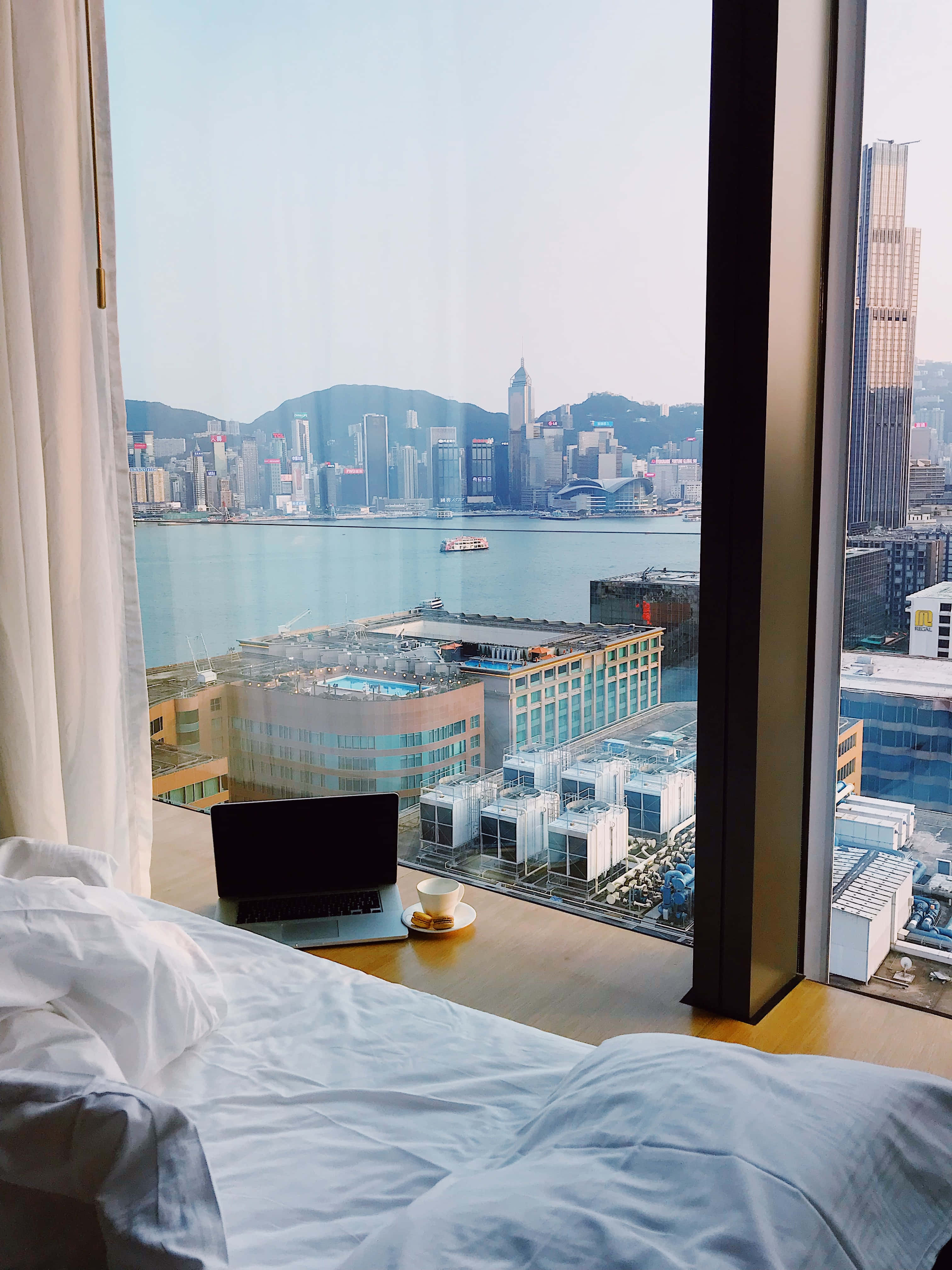 Hotel Room 桌布 壁紙 — 舒適的飯店房間，在木製窗台上放著筆記型電腦和咖啡，俯瞰香港的天際線和港口。