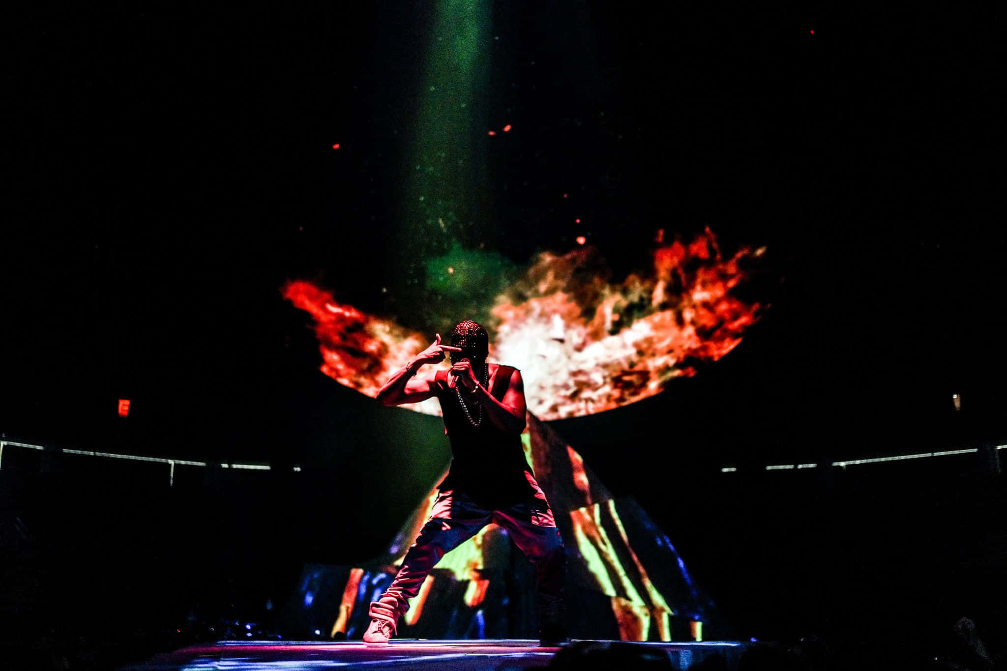Kanye West Yeezus 桌布 壁紙 — 舞台上的表演者在黑暗的音樂會場景中，以火焰翅膀和綠色燈光襯托出剪影。
