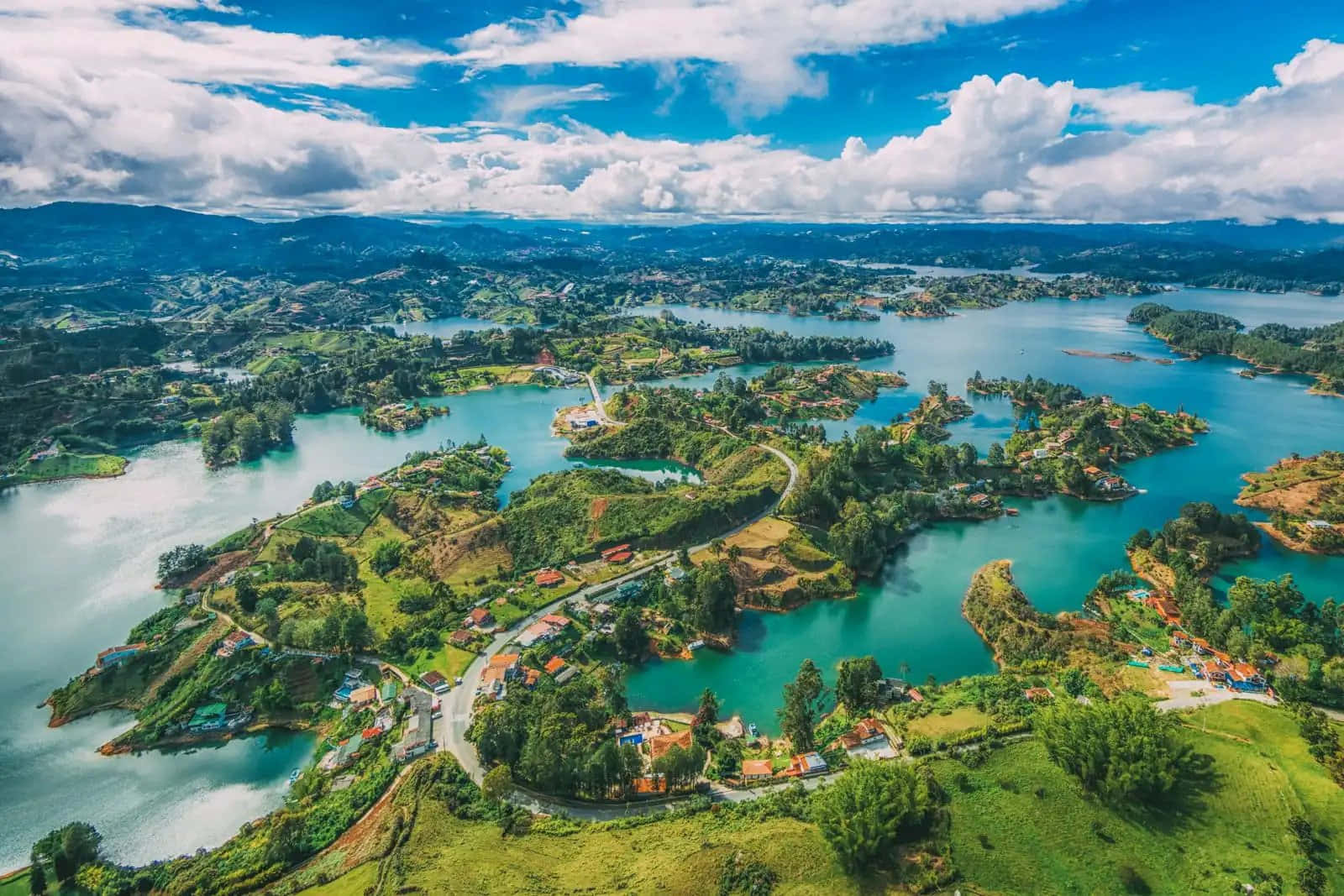 Colombia 桌布 壁紙 — 航拍視角，碧綠的湖泊蜿蜒穿過鬱鬱蔥蔥的綠色島嶼，島上點綴著色彩繽紛的房屋，上方是明亮的藍天和蓬鬆的白雲。