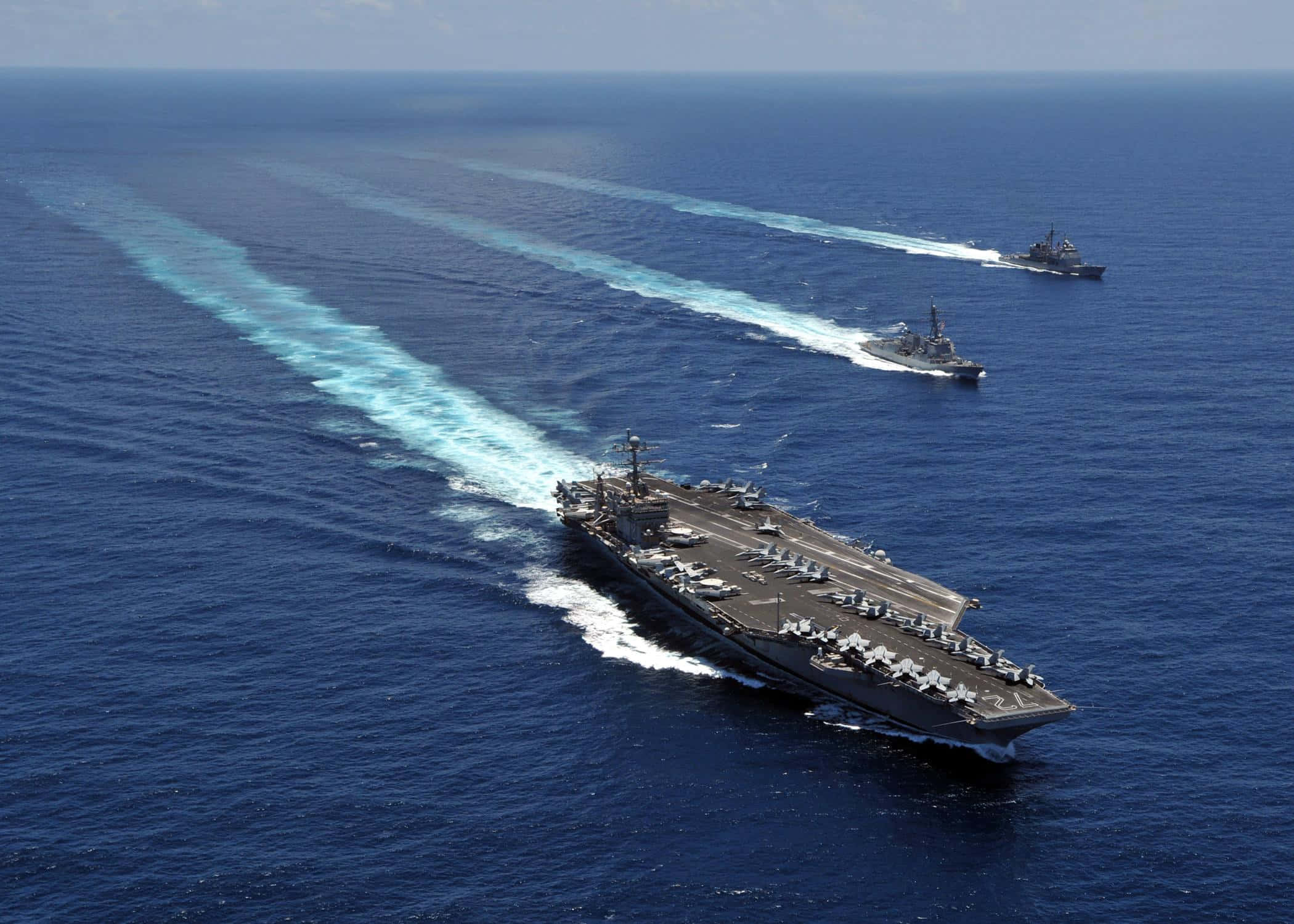 Navy 桌布 壁紙 — 航空母艦和兩艘驅逐艦在晴朗天空下劃過深藍色海洋，留下白色航跡。