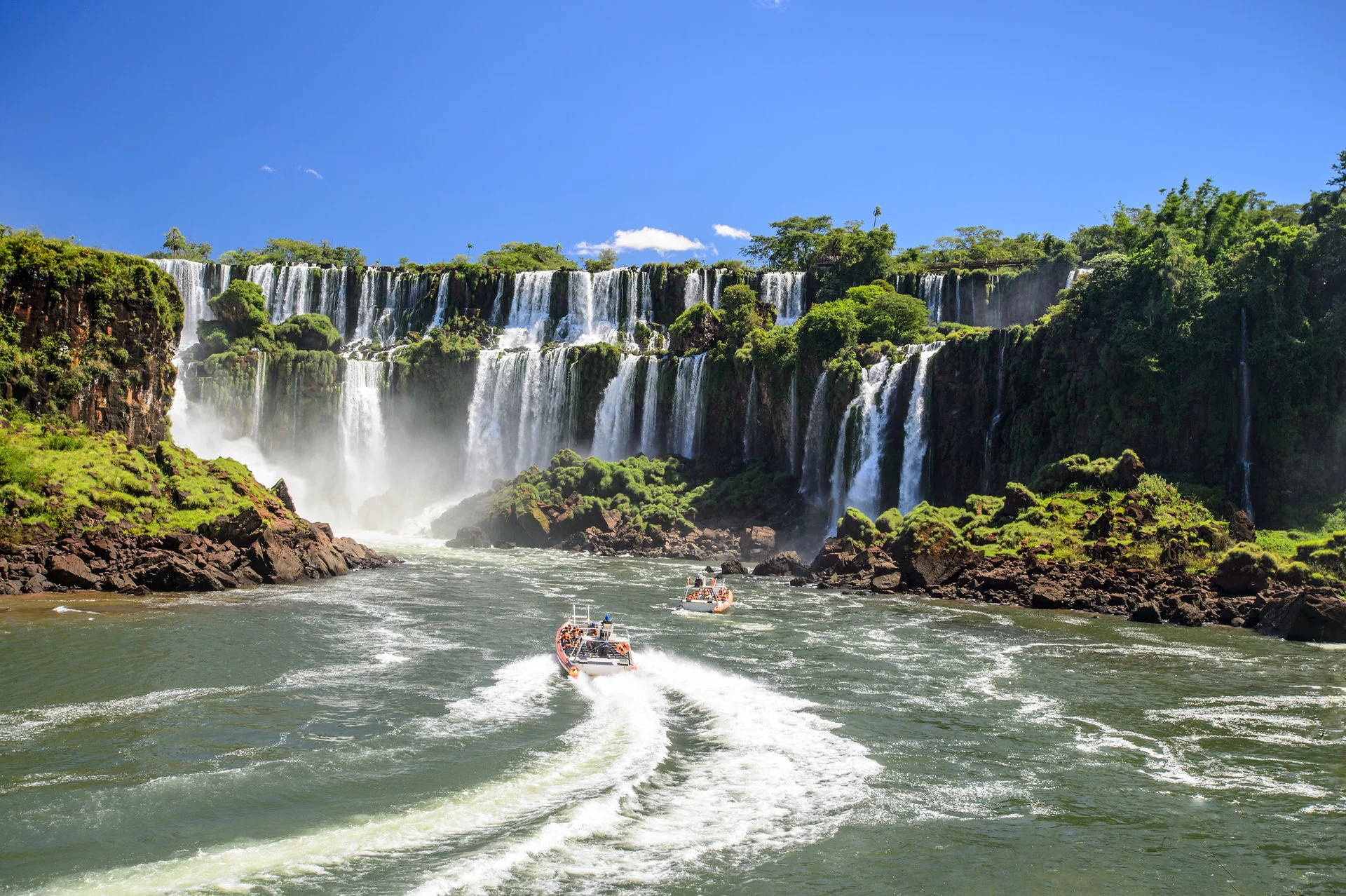 South America 桌布 壁紙 — 船隻在轟鳴的伊瓜蘇瀑布下巡航，瀑布水流與茂密的綠色懸崖相遇，天空清澈湛藍。