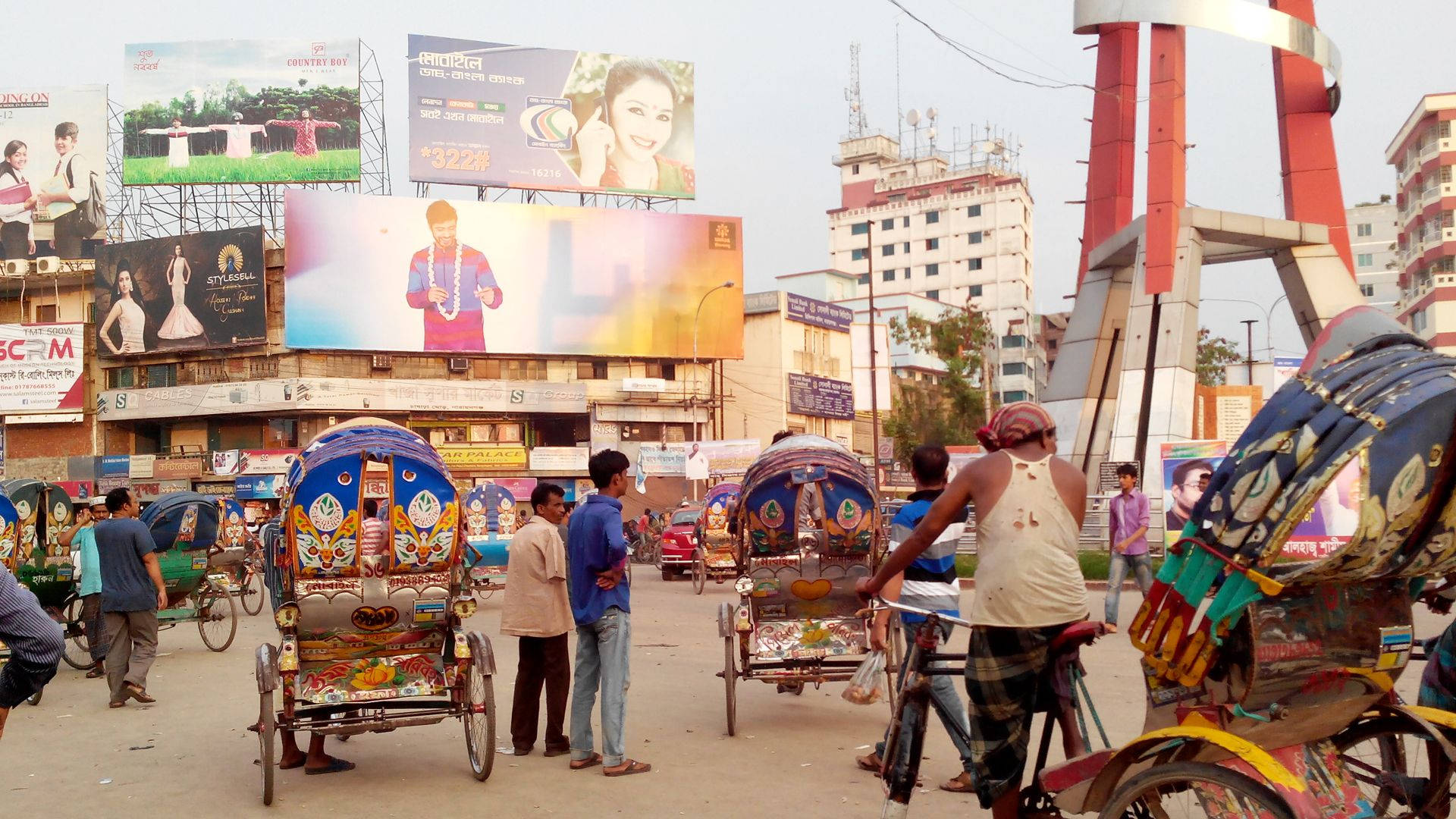 Bangladesh 桌布 壁紙 — 色彩繽紛、裝飾華麗的人力車在繁忙的城市街道上行駛，街道兩旁是高聳的廣告牌和現代建築，天空朦朧。