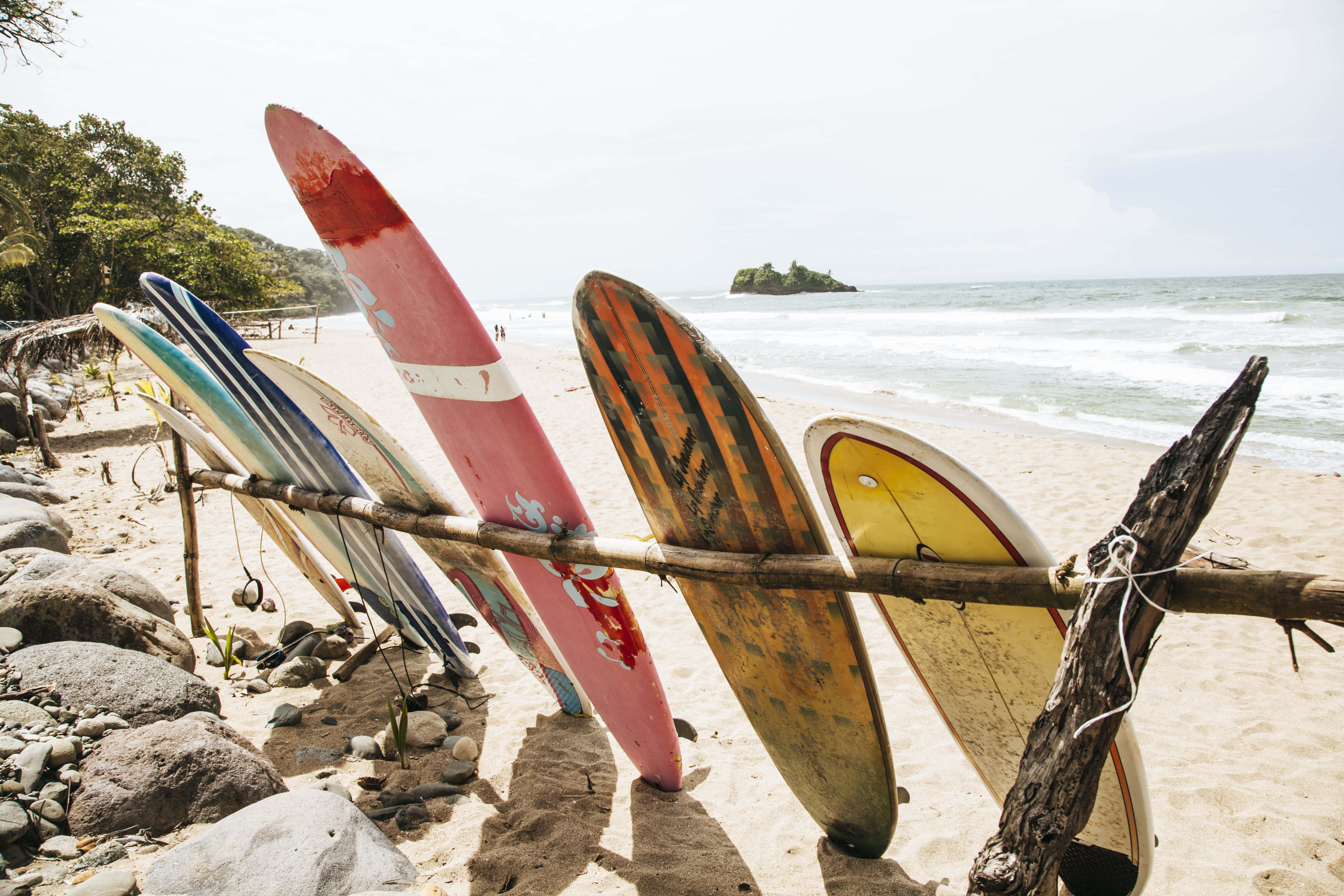 surfing 桌布 壁紙 — 色彩繽紛、飽經風霜的衝浪板靠在浮木架上，位於陽光明媚的沙灘上，有海浪和遠處的島嶼。