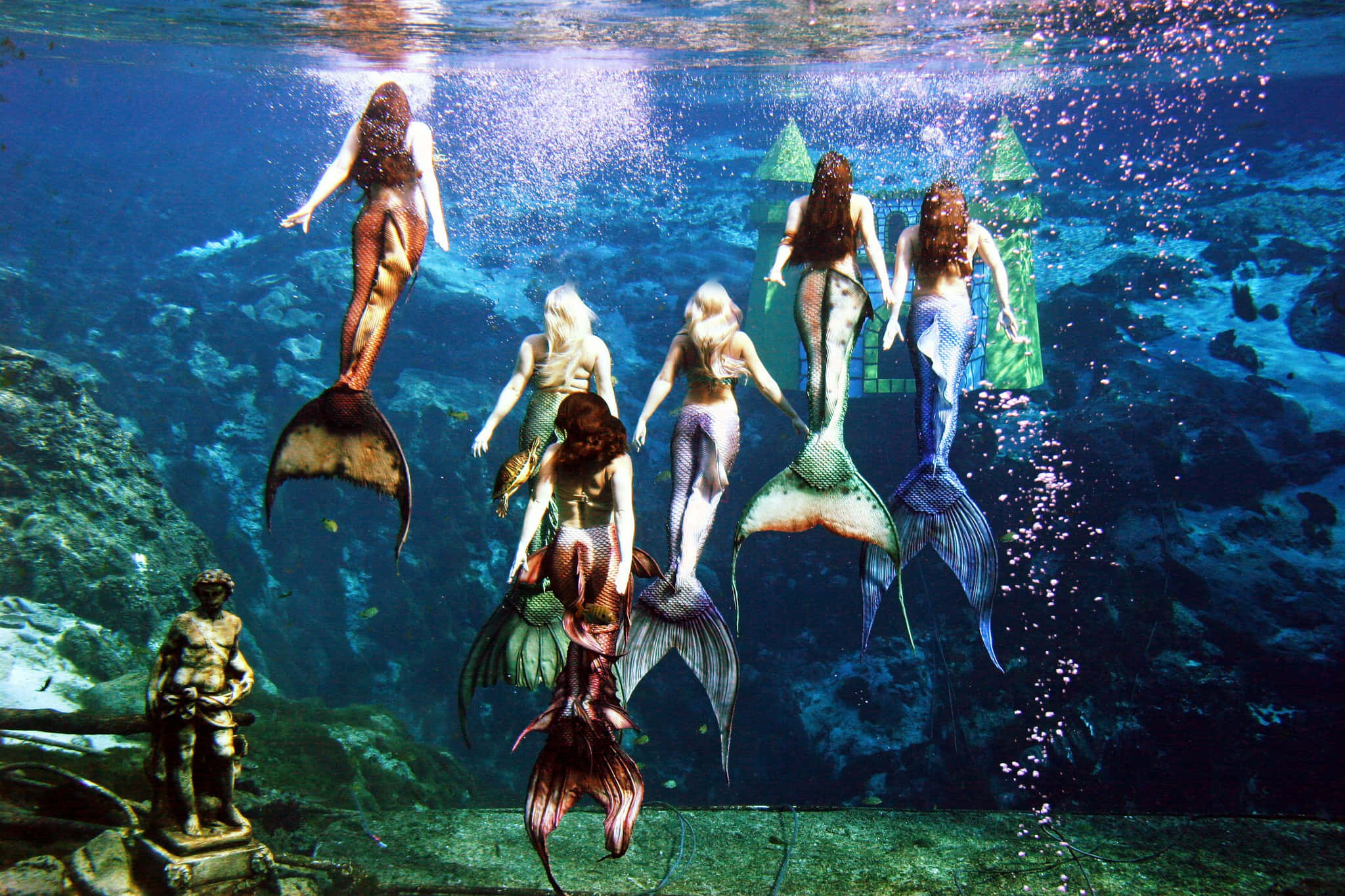 Real Mermaid 桌布 壁紙 — 色彩繽紛尾巴的美人魚在 submerged 雕像和綠色瓷磚結構附近優雅地滑行。