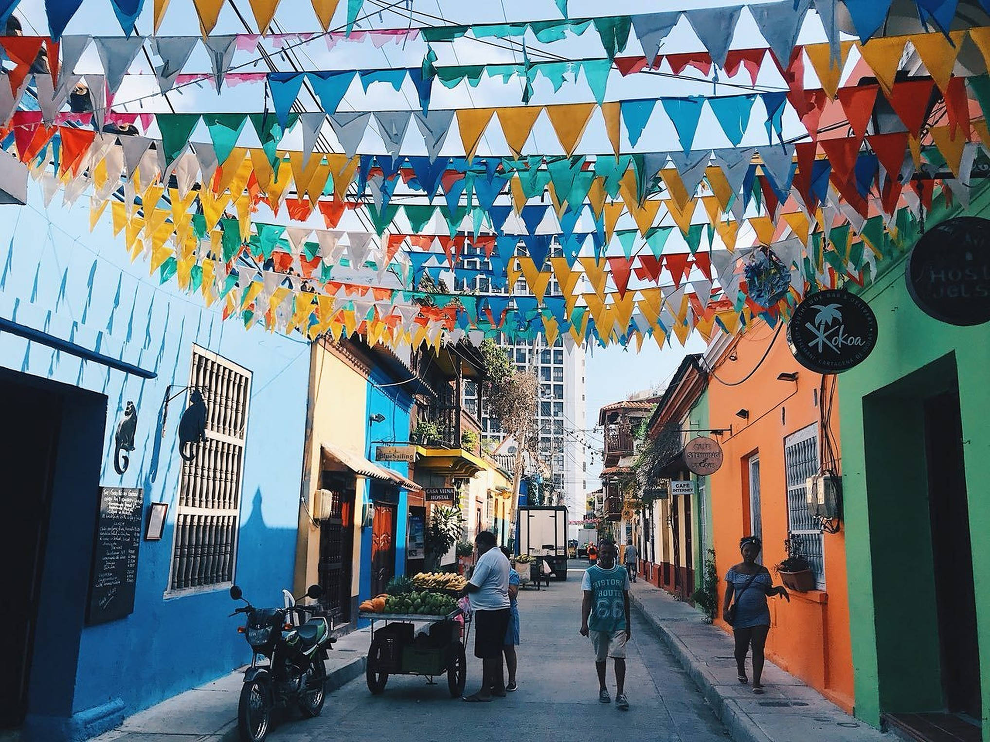 Colombia 桌布 壁紙 — 色彩繽紛的三角形旗幟懸掛在一條熱鬧狹窄的街道上方，街道兩旁是明亮的藍色、橙色和綠色建築物。