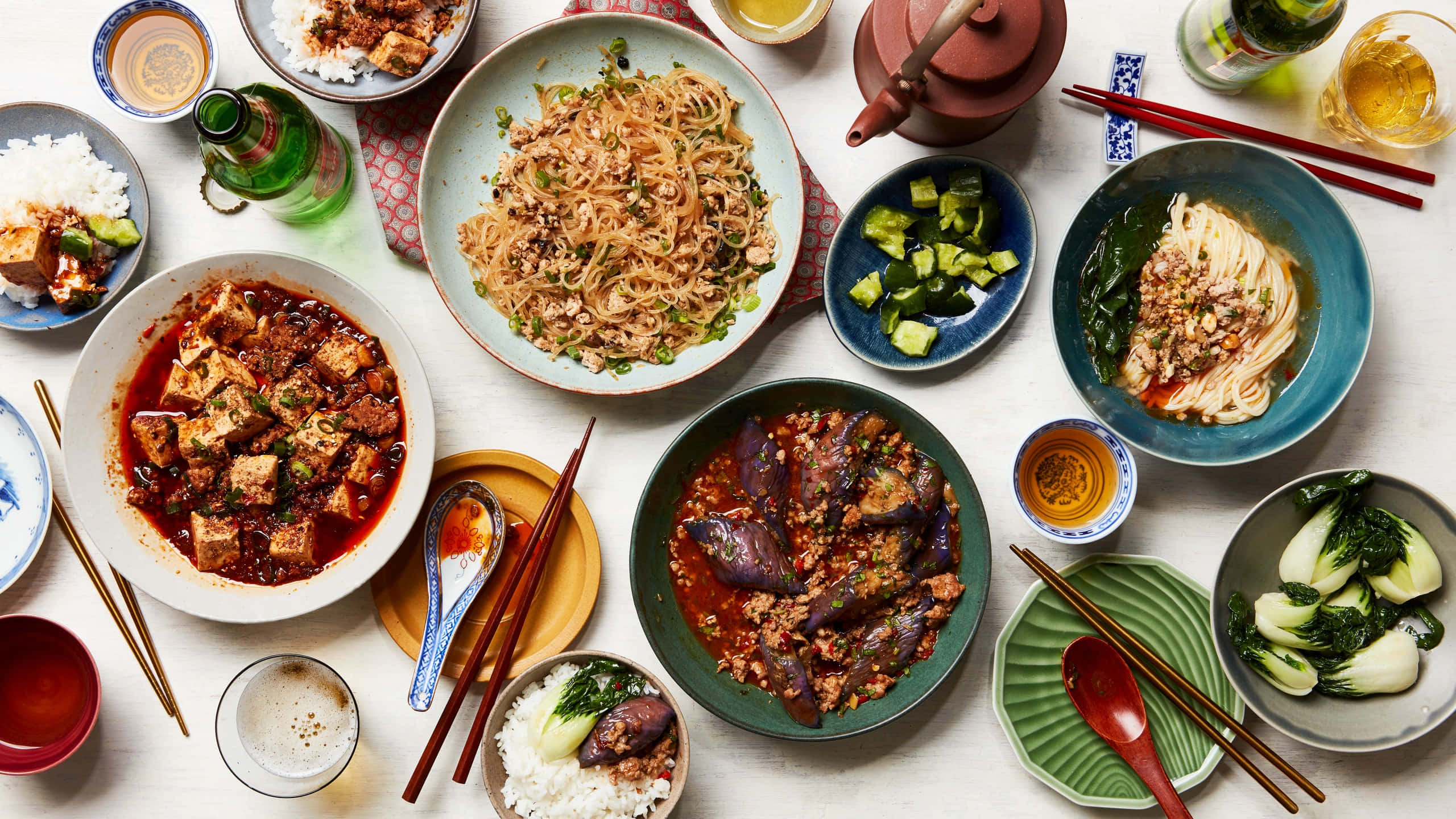 Food Table 桌布 壁紙 — 色彩繽紛的亞洲菜餚俯視圖，包括麵條、豆腐和茄子，盛在陶瓷碗中，附有筷子和飲料。