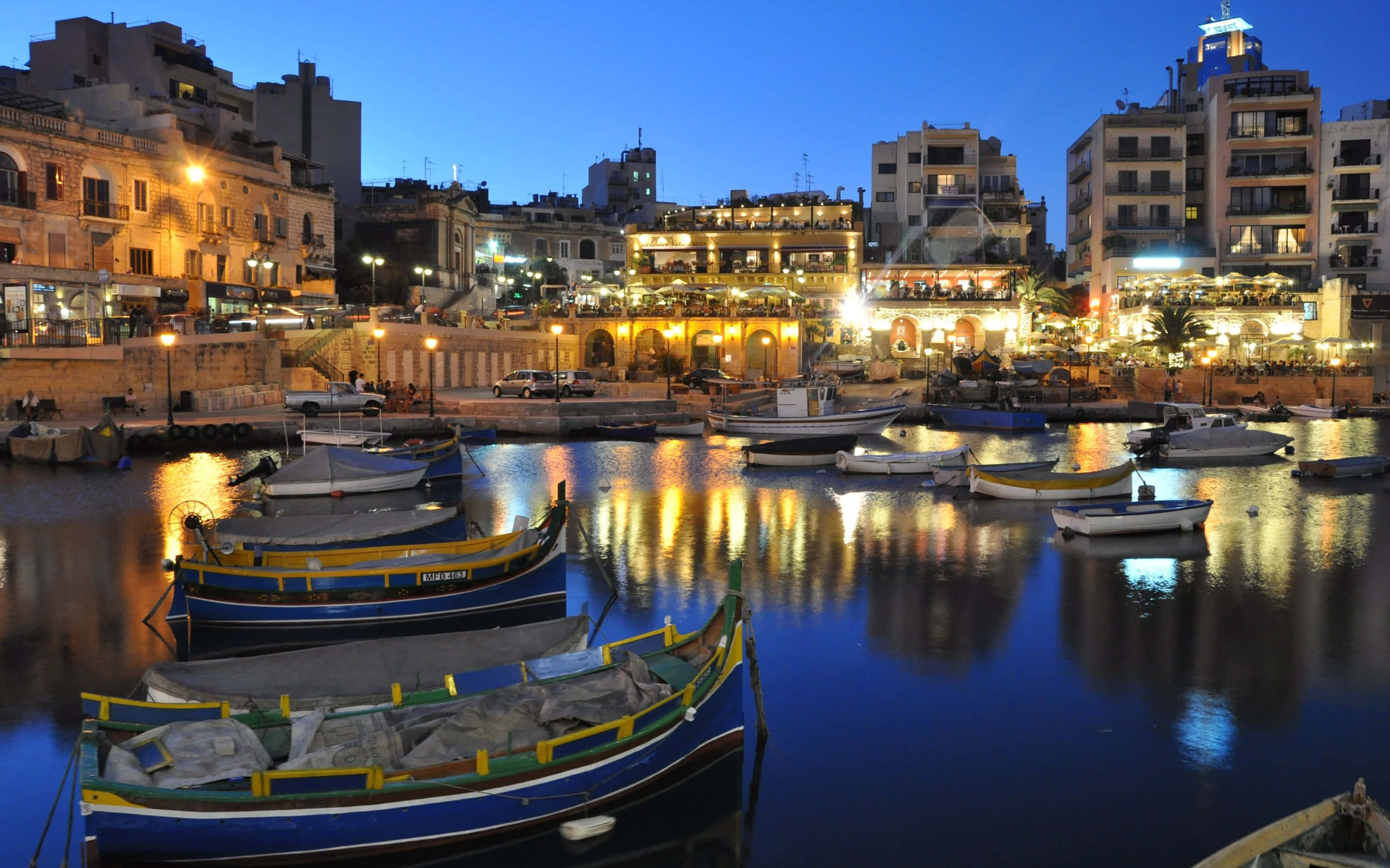 Malta 桌布 壁紙 — 色彩繽紛的傳統馬耳他船隻在黃昏時分停泊在寧靜的港口，歷史建築的溫暖燈光倒映在平靜的水面上。
