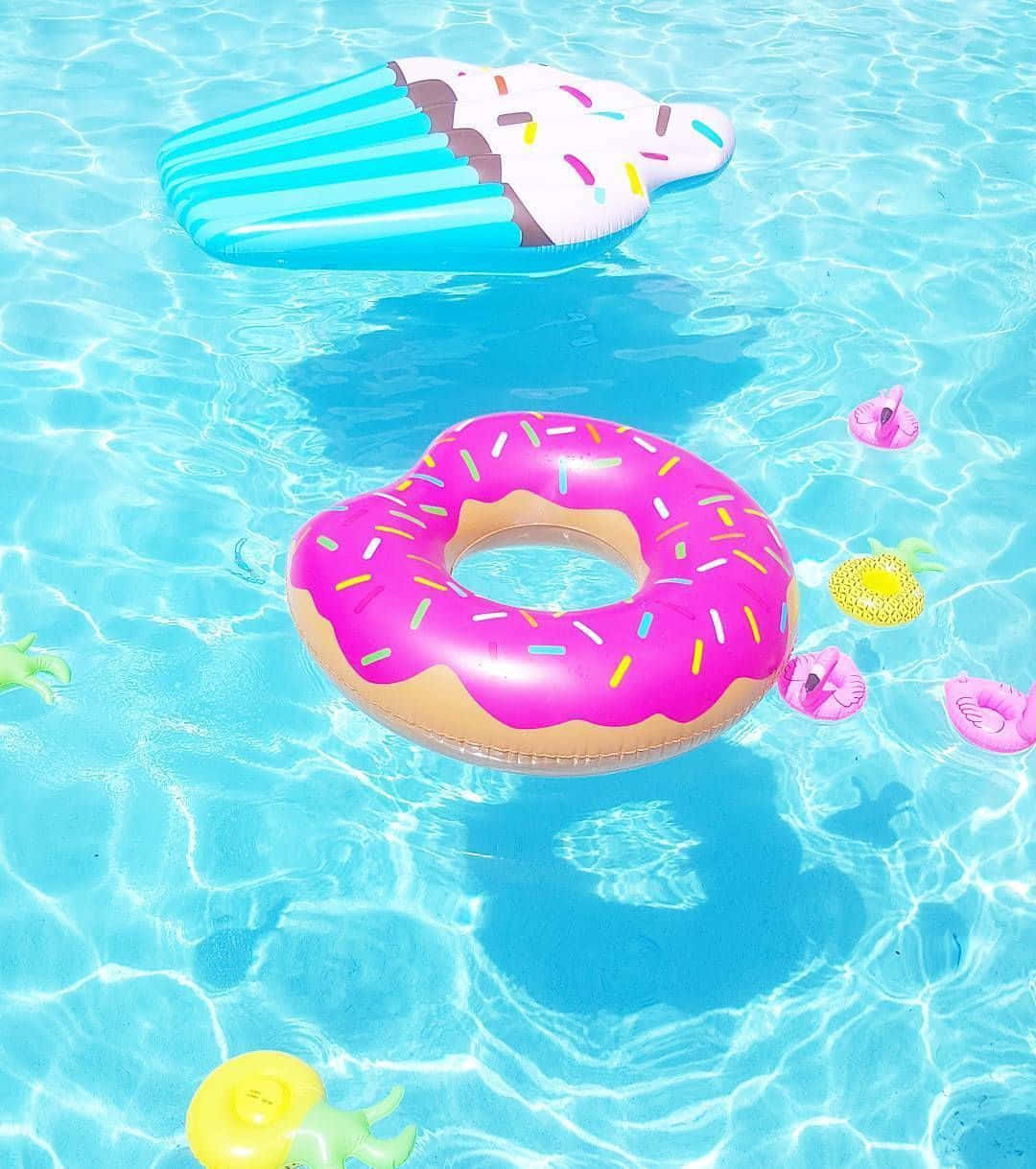 Cupcake 桌布 壁紙 — 色彩繽紛的充氣甜甜圈和冰淇淋浮床在閃爍的藍綠色泳池水中與有趣的玩具一起漂浮。