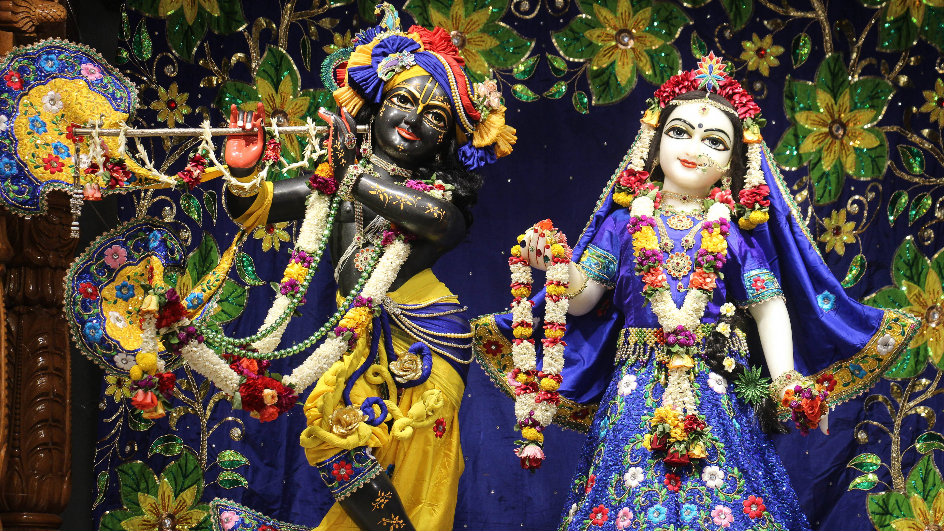 krishna ji 桌布 壁紙 — 色彩繽紛的克里希納和拉達雕像，飾以鮮花和珠寶，背景為花卉掛毯。
