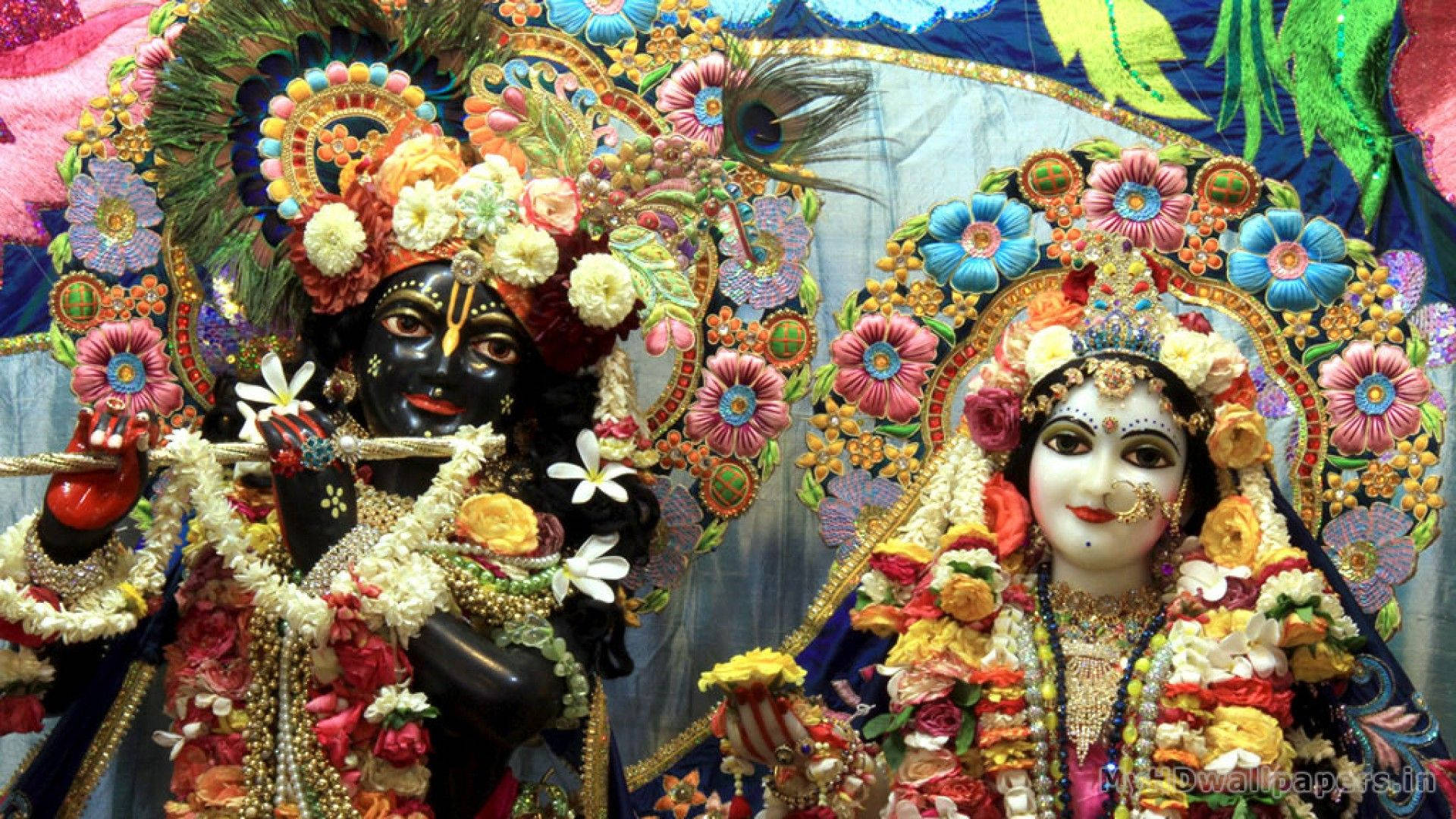 radha krishna HD 桌布 壁紙 — 色彩繽紛的印度教神祇克里希納和拉妲並肩而立，頭戴花冠、身披花環，背景圖案繁複。