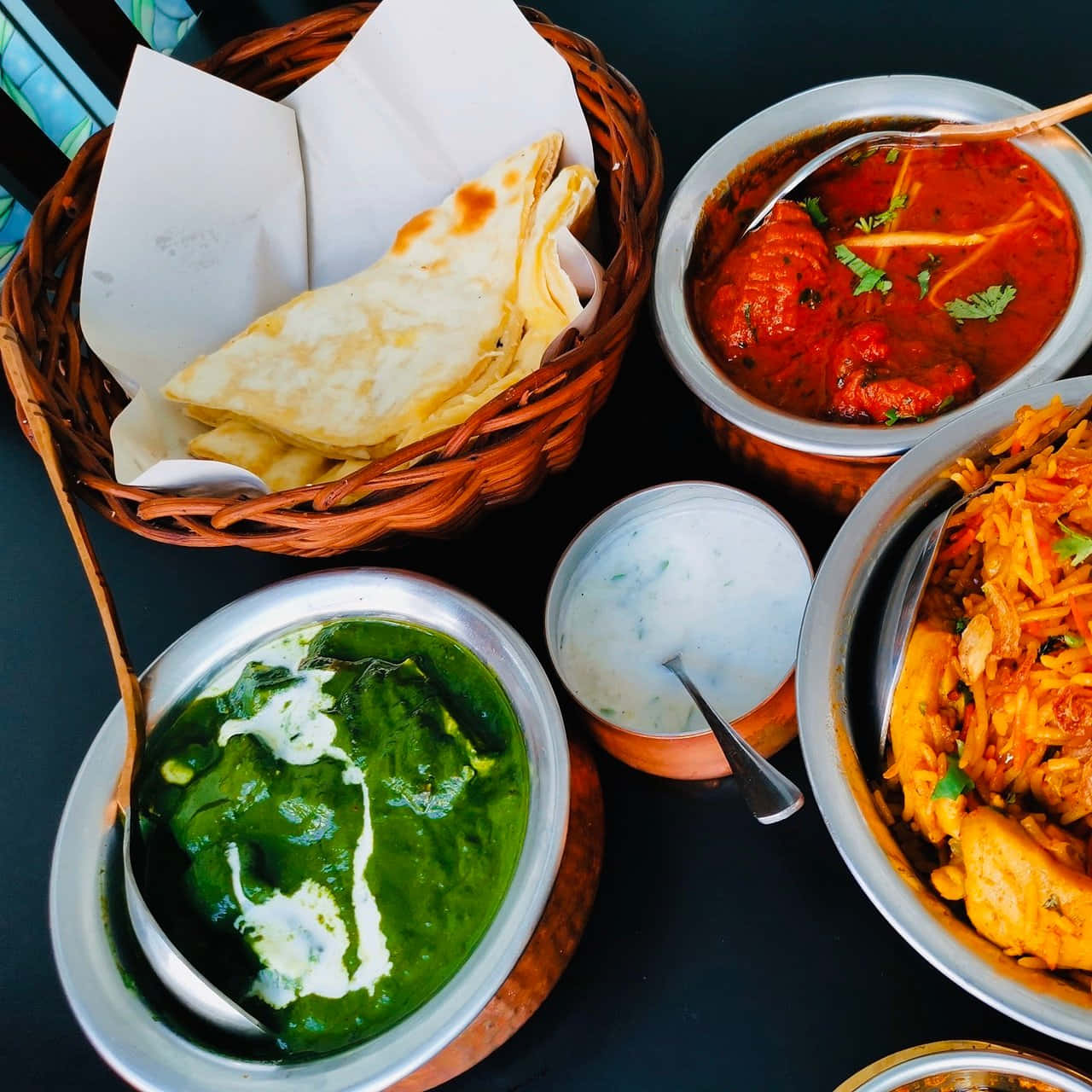 Indian 桌布 壁紙 — 色彩繽紛的印度菜餚，包括菠菜咖哩、番茄咖哩、米飯和烤餅，盛放在金屬碗和編織籃中。