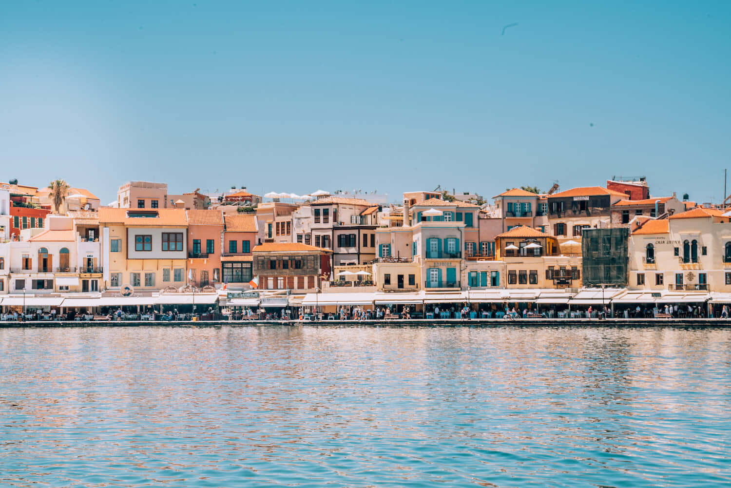 Greek Island 桌布 壁紙 — 色彩繽紛的地中海建築沿著熱鬧的海濱排列，天空清澈湛藍，水面泛起輕柔的漣漪。