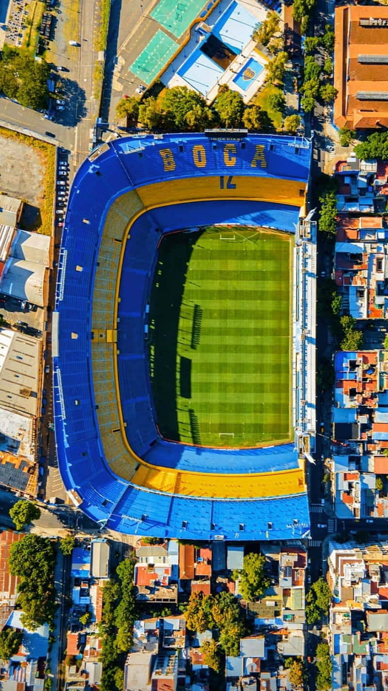 Stadium 桌布 壁紙 — 色彩繽紛的城市屋頂中，一個充滿活力的藍黃色足球場的鳥瞰圖。