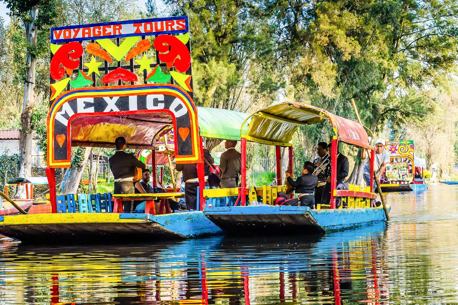 墨西哥 桌布 壁紙 — 色彩繽紛的墨西哥特拉希內拉船隻帶有「Mexico」標誌，在陽光明媚的日子裡，在枝葉茂密的樹木下滑過平靜的水面。