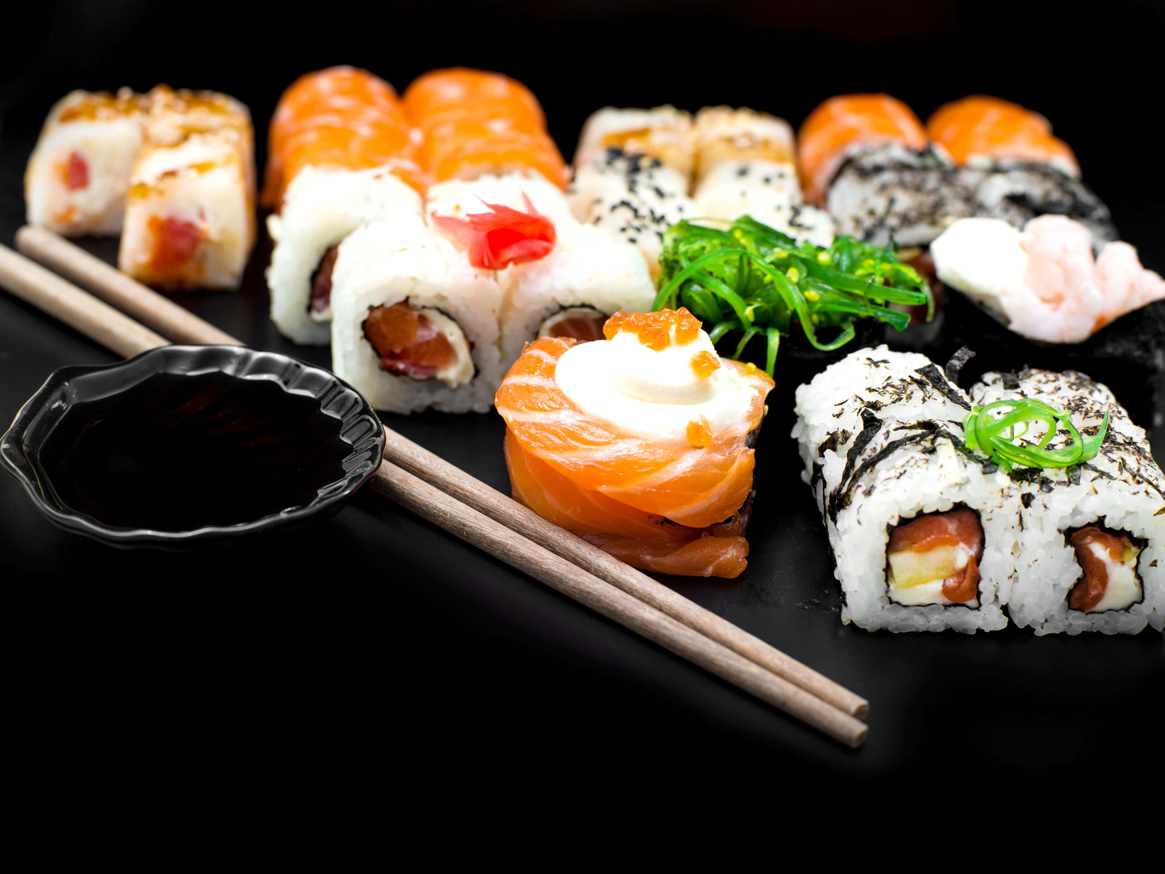 Sushi 桌布 壁紙 — 色彩繽紛的壽司卷和握壽司，藝術性地擺放在黑色盤子上，配上醬油和筷子。