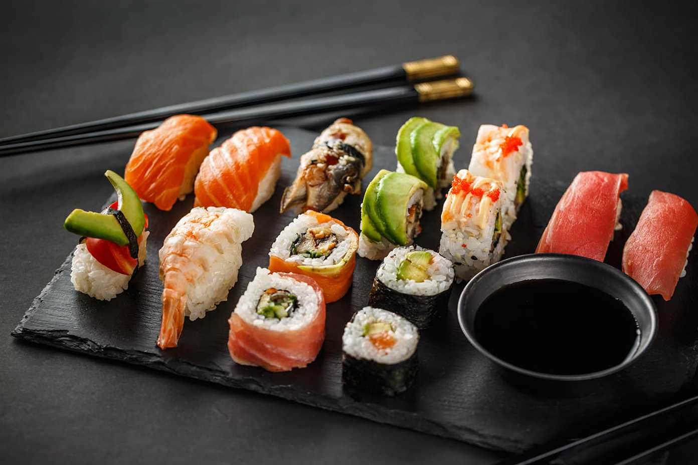 Sushi 桌布 壁紙 — 色彩繽紛的壽司拼盤，藝術性地擺放在深色石板盤上，搭配醬油和筷子，令人垂涎。