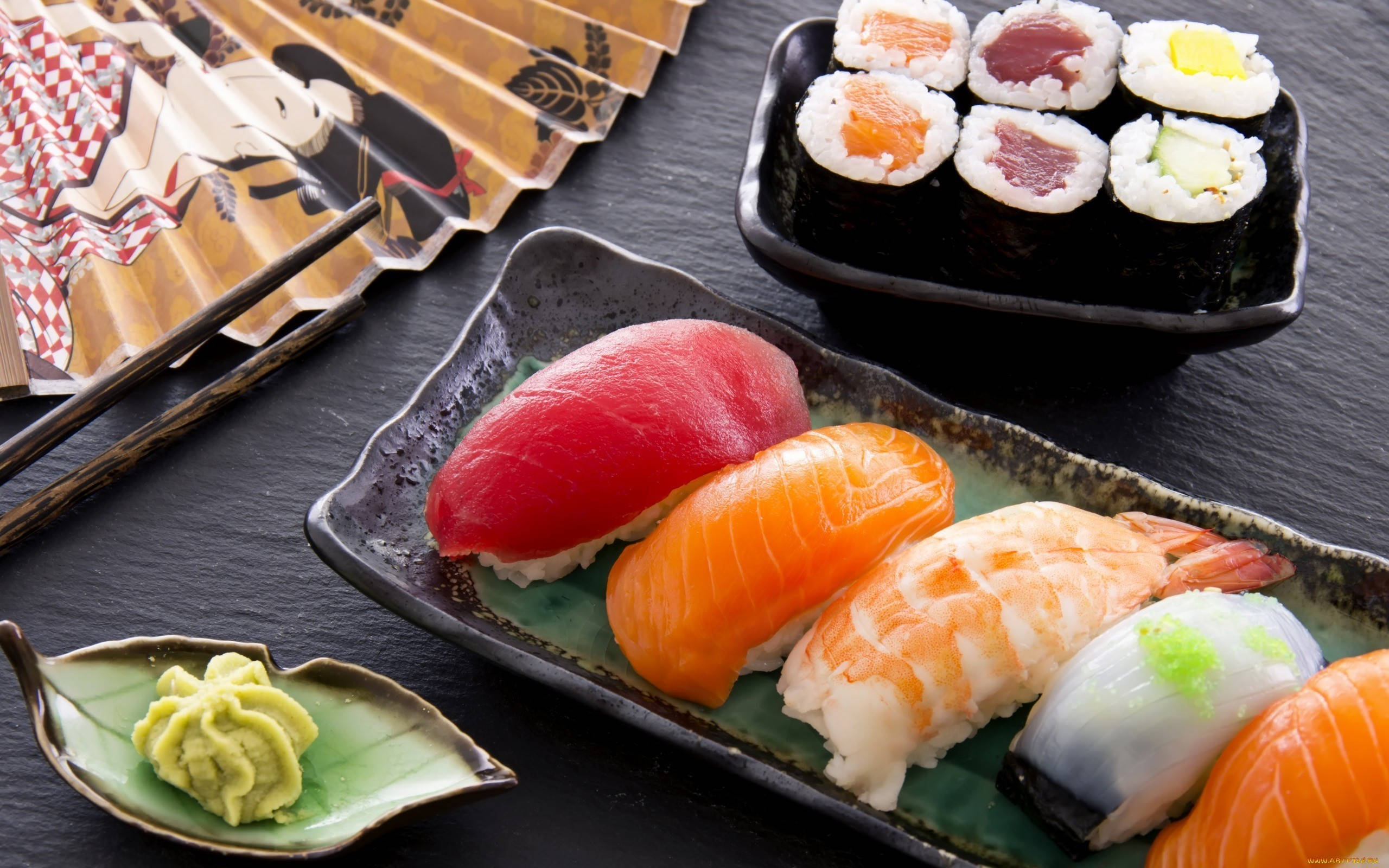 Sushi 桌布 壁紙 — 色彩繽紛的壽司拼盤近照，包括鮪魚、鮭魚和蝦，擺放在深色陶瓷盤上，旁邊有芥末和筷子。