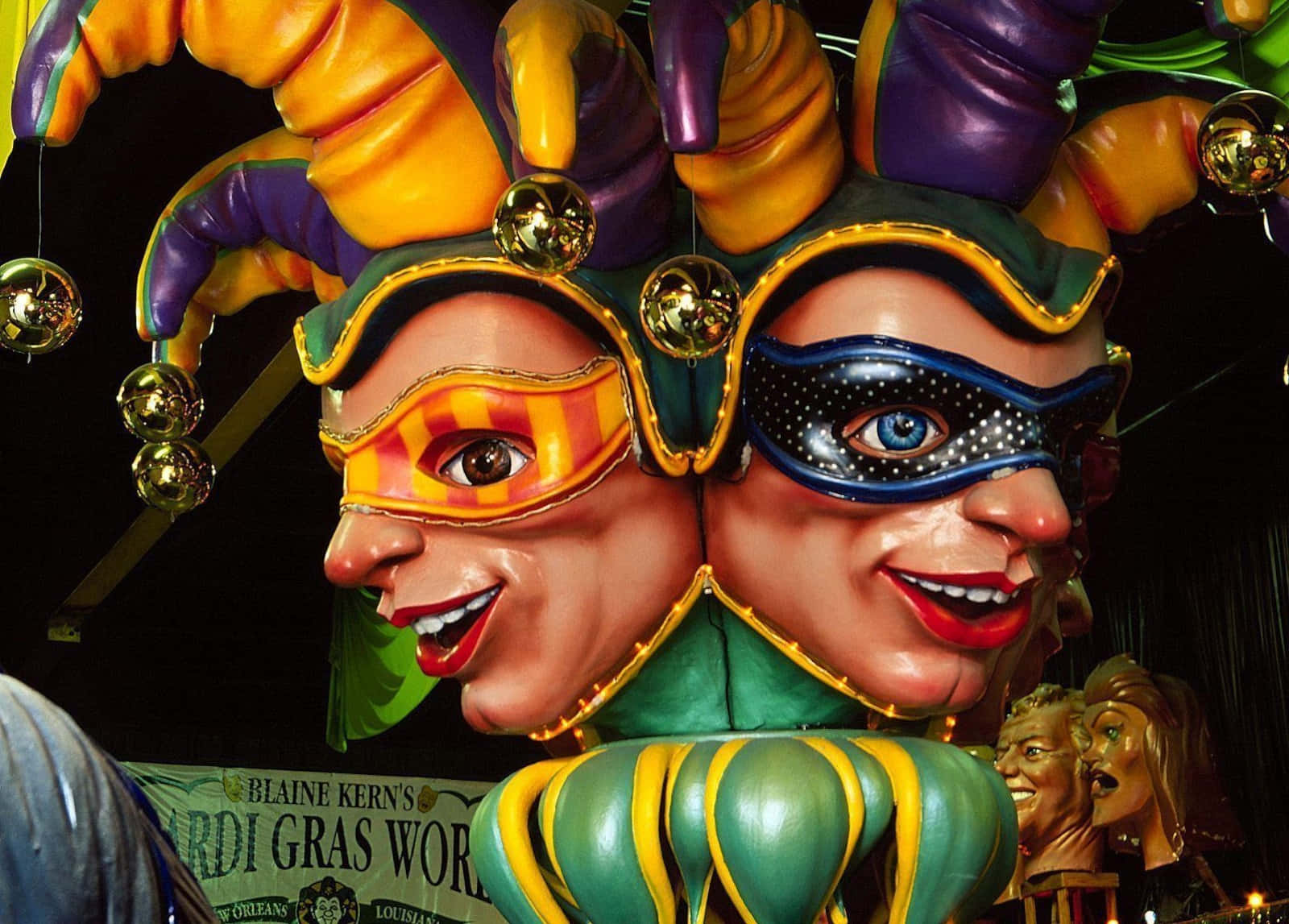 Mardi Gras 桌布 壁紙 — 色彩繽紛的小丑面具，帶有彩色角和閃閃發光的鈴鐺，在夜燈下裝飾著節日的Mardi Gras花車。