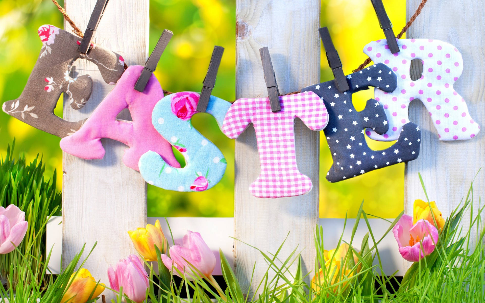 Easter 桌布 壁紙 — 色彩繽紛的布料字母拼出 'EASTER'，掛在木柵欄上，下方是盛開的粉紅和黃色鬱金香，點綴在充滿活力的春季草地上。