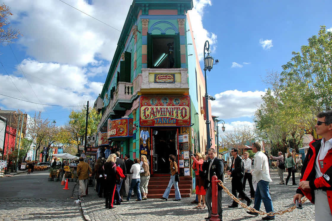 Argentina 桌布 壁紙 — 色彩繽紛的建築物帶有「Caminito Tango」標誌，在陽光明媚、綠樹成蔭的鵝卵石街道上吸引人群。