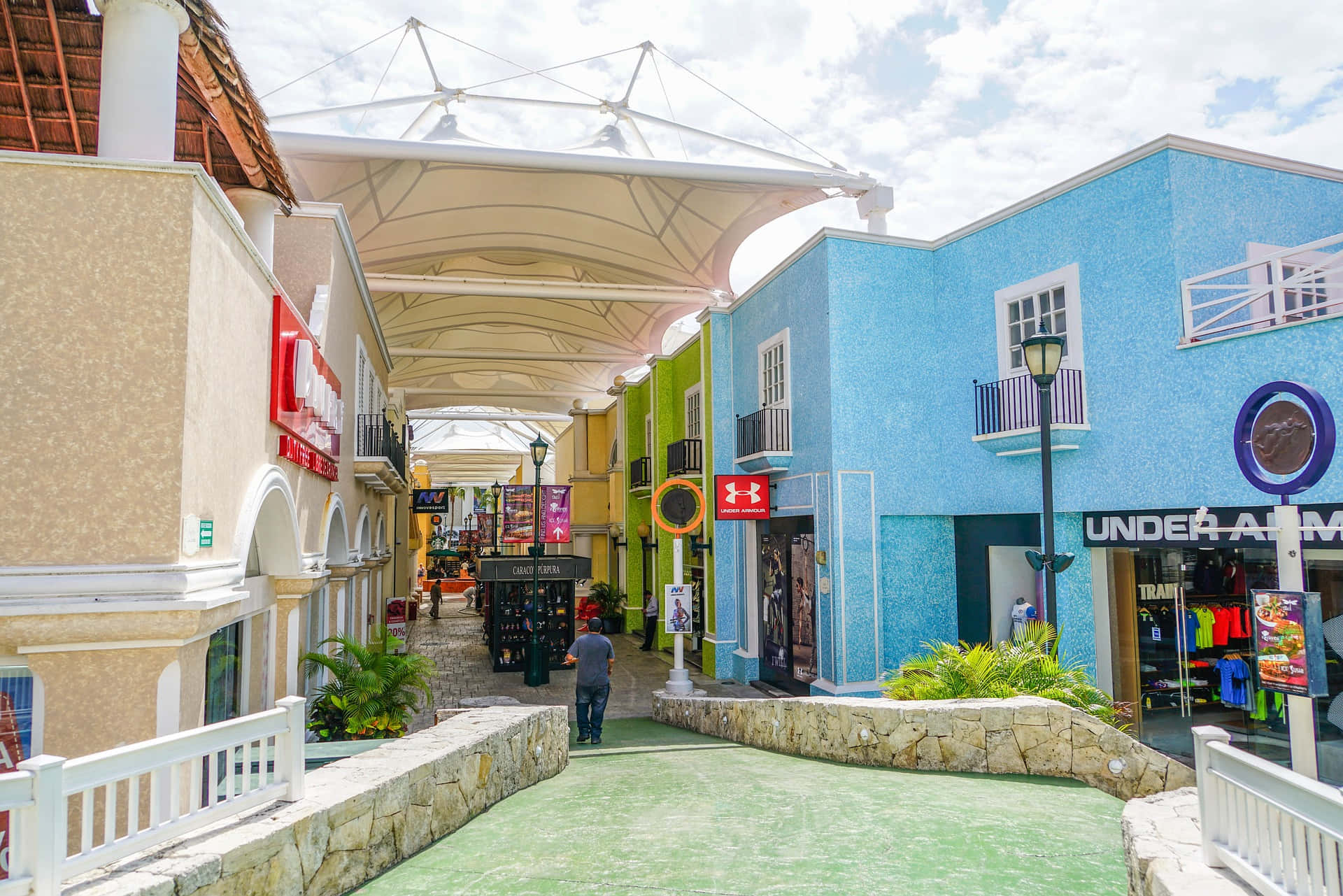 Cancun 桌布 壁紙 — 色彩繽紛的建築物排列在一條有遮蔭的行人通道上，通道有綠色地板和石牆，上方是明亮的天空。