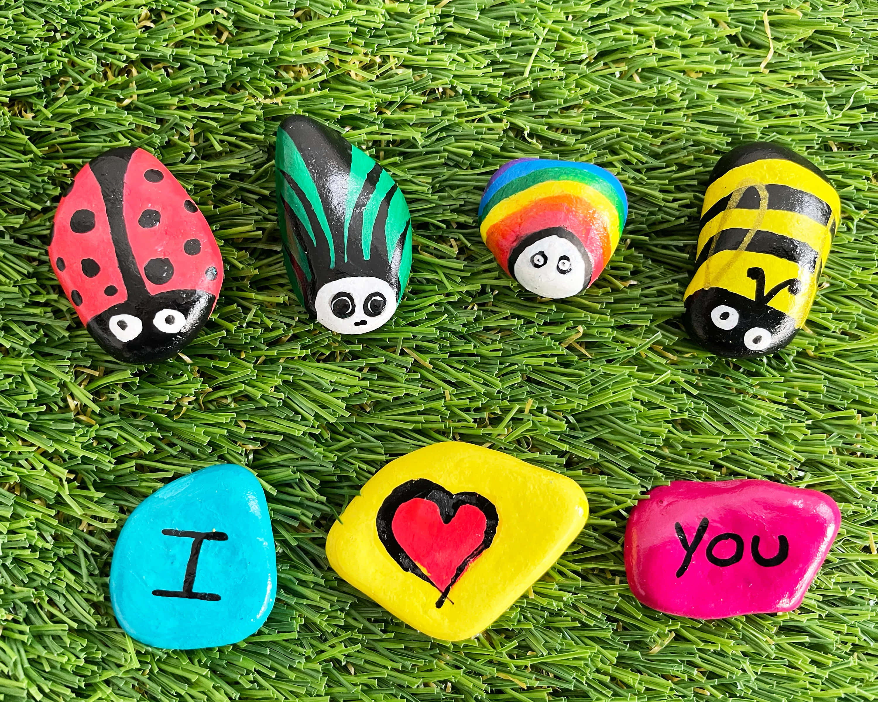 Pebble Art 桌布 壁紙 — 色彩繽紛的彩繪石頭，形狀像瓢蟲、蜜蜂和心形，拼出「我愛你」，放在充滿活力的綠色草地上。
