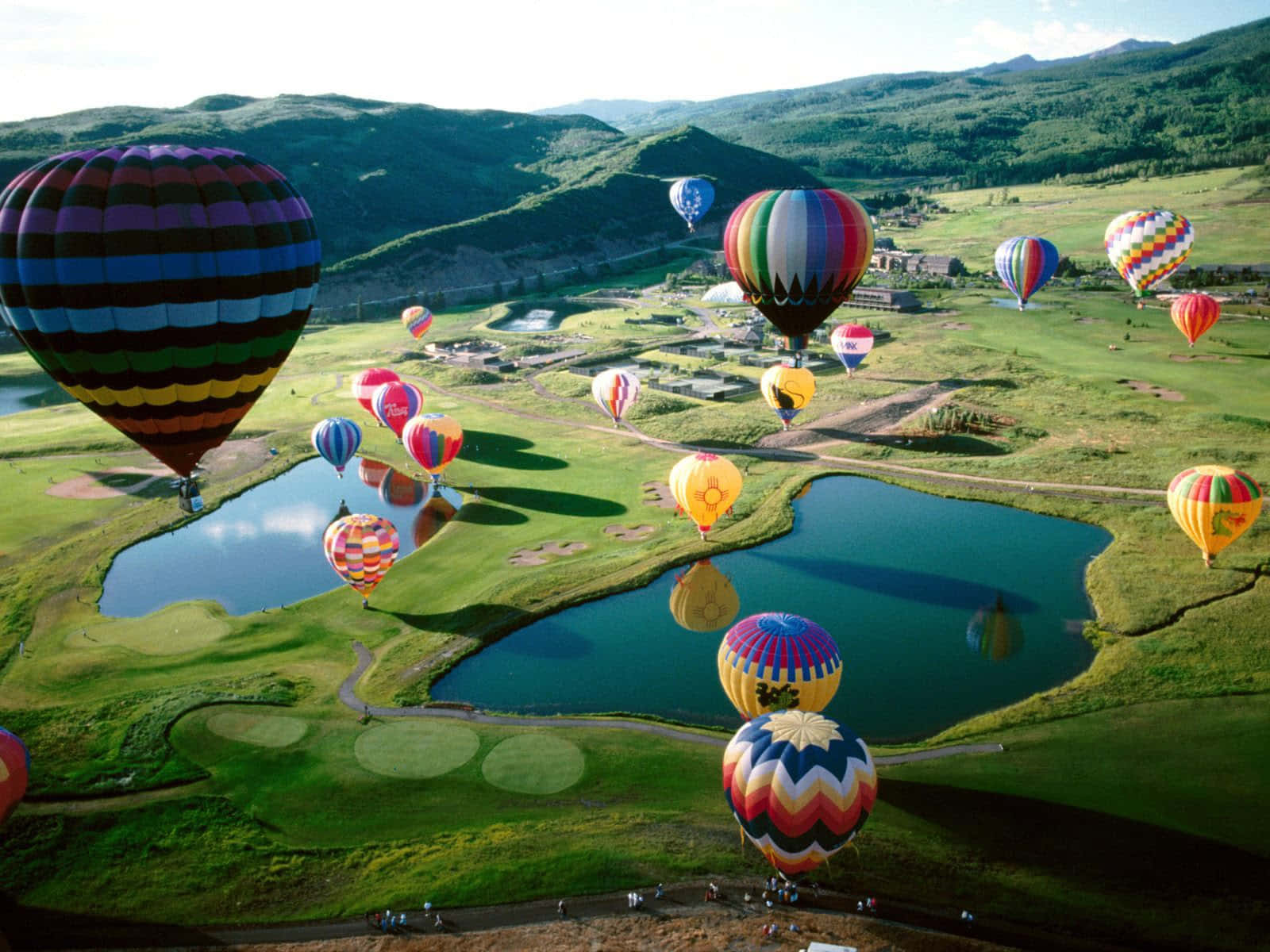 Hot Air Balloon 桌布 壁紙 — 色彩繽紛的彩虹圖案熱氣球在明亮的日光下，飄浮在一個擁有蜿蜒湖泊和起伏山丘的茂密綠色高爾夫球場上空。