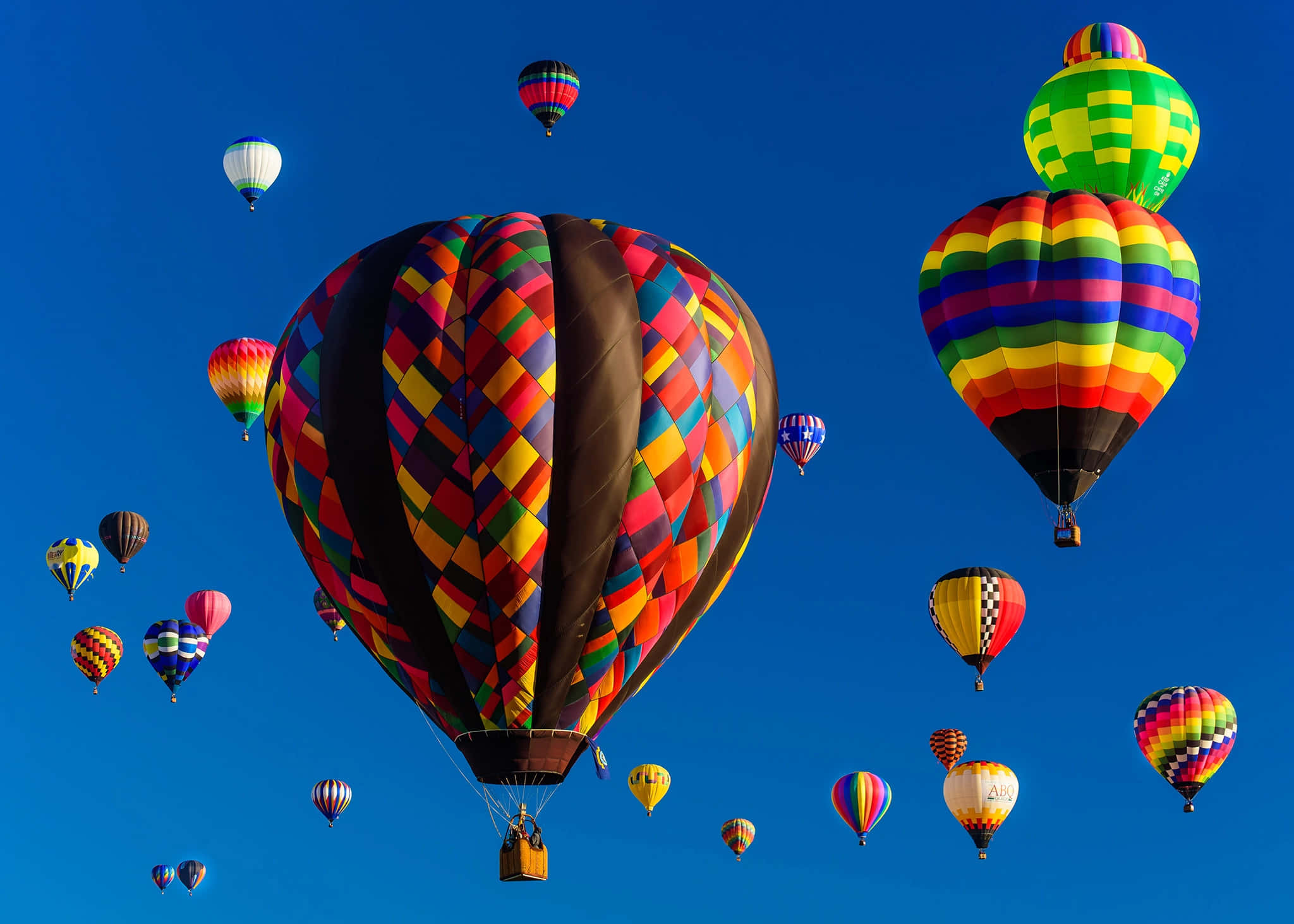 Hot Air Balloon 桌布 壁紙 — 色彩繽紛的彩虹圖案熱氣球在晴朗的藍天中飄浮，構成節日氣氛的空中聚會。