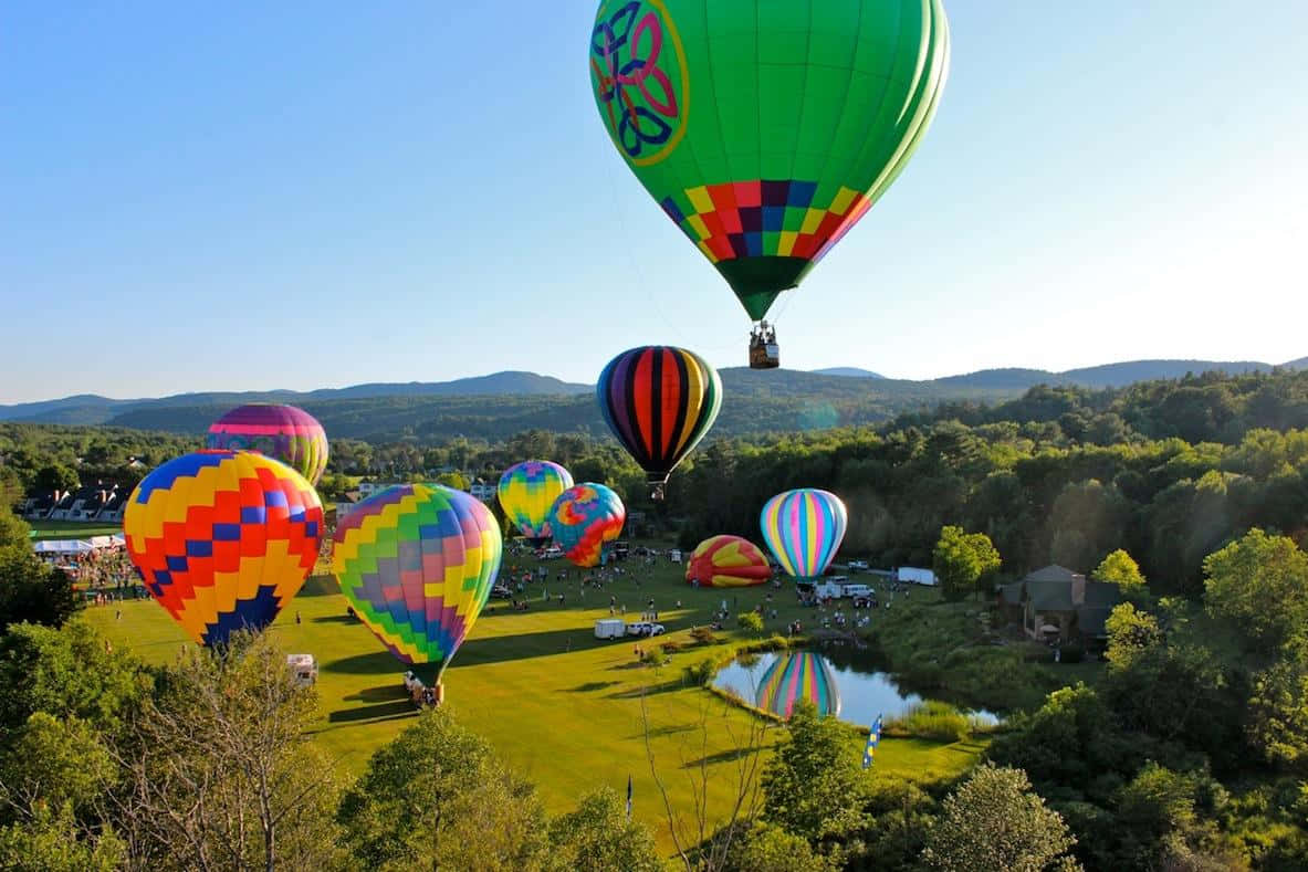 Hot Air Balloon 桌布 壁紙 — 色彩繽紛的彩虹圖案熱氣球在晴朗藍天下，漂浮並停歇在池塘附近、林木茂密的山丘旁一片翠綠的田野上。