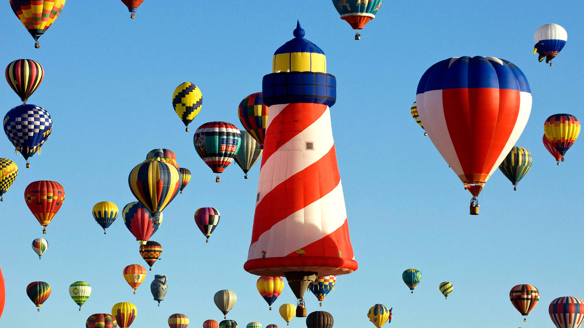 Hot Air Balloon 桌布 壁紙 — 色彩繽紛的彩虹圖案熱氣球在藍天白雲下圍繞著紅白條紋燈塔飄浮。