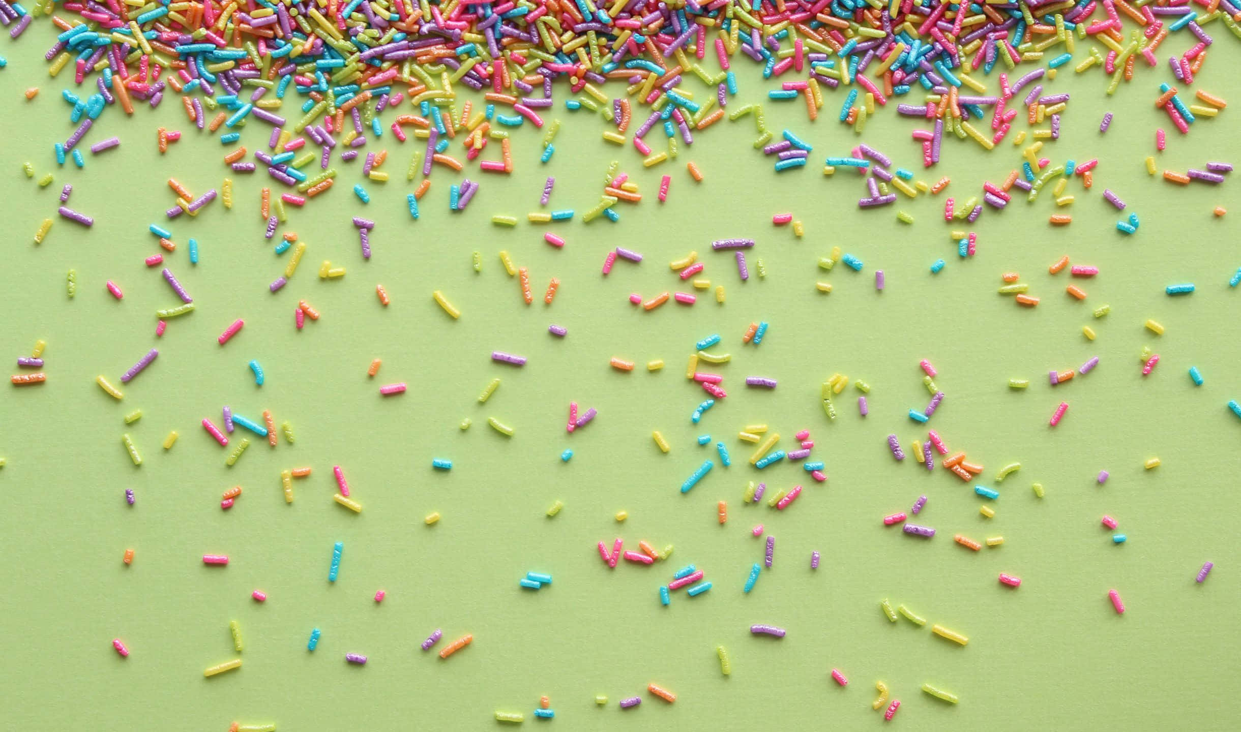 Sprinkles 桌布 壁紙 — 色彩繽紛的彩虹糖針在平滑的淺綠色表面上以對角線方式傾瀉而下，營造出俏皮的俯拍效果。
