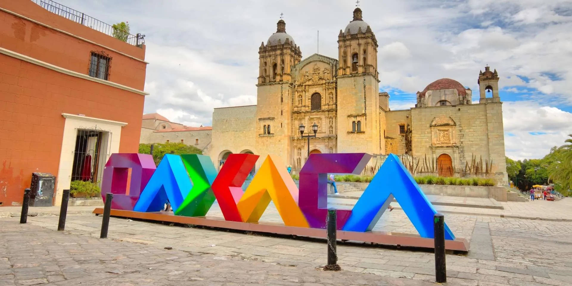 墨西哥 桌布 壁紙 — 色彩繽紛的彩虹色「OAXACA」字母，矗立在歷史悠久的石砌大教堂前的鵝卵石廣場上，天空部分多雲。