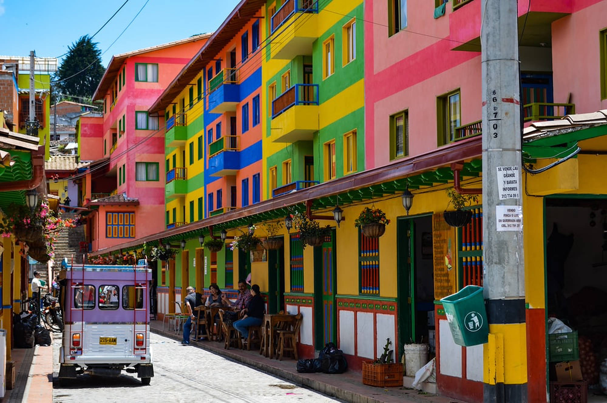 Colombia 桌布 壁紙 — 色彩繽紛的彩虹色建築物排列在一條熱鬧的鵝卵石街道上，人們在晴朗的天空下在戶外用餐。