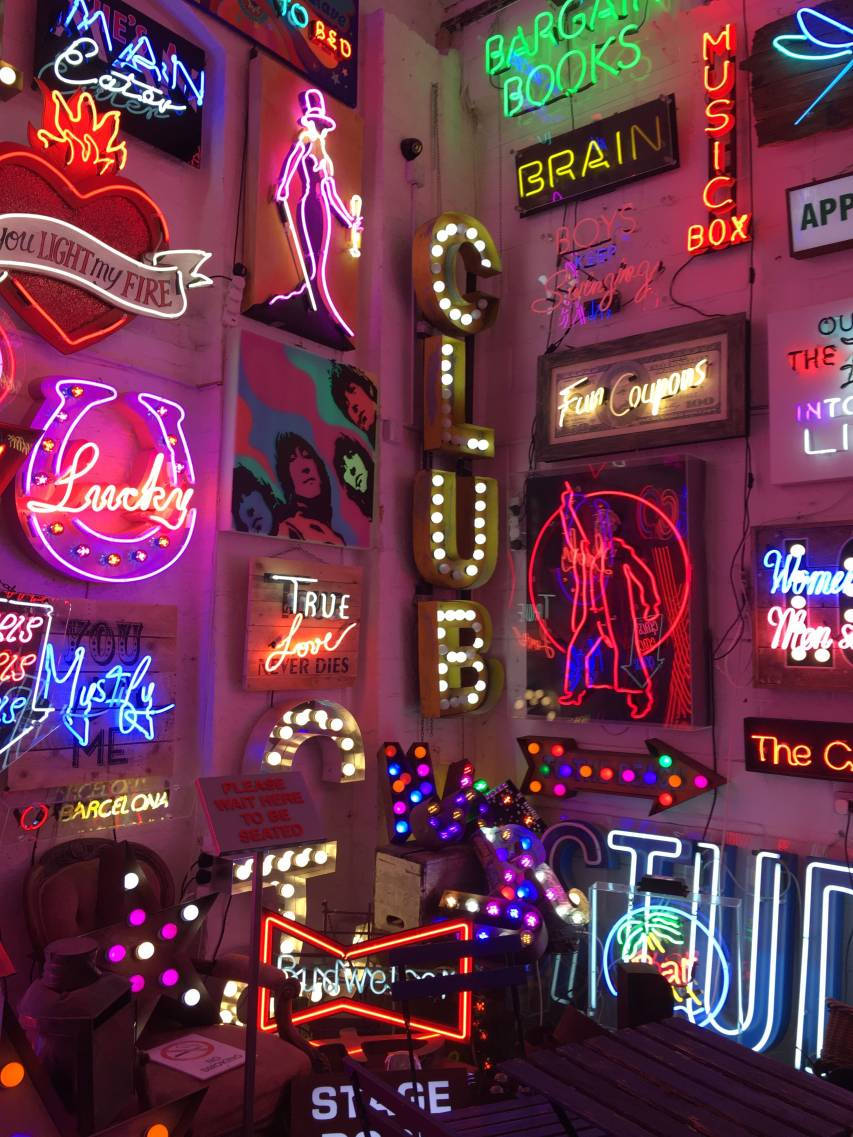 dark neon iPhone 桌布 壁紙 — 色彩繽紛的復古字體中，密集的牆上展示著俱樂部、書籍和音樂，鮮豔的霓虹燈招牌閃閃發光。