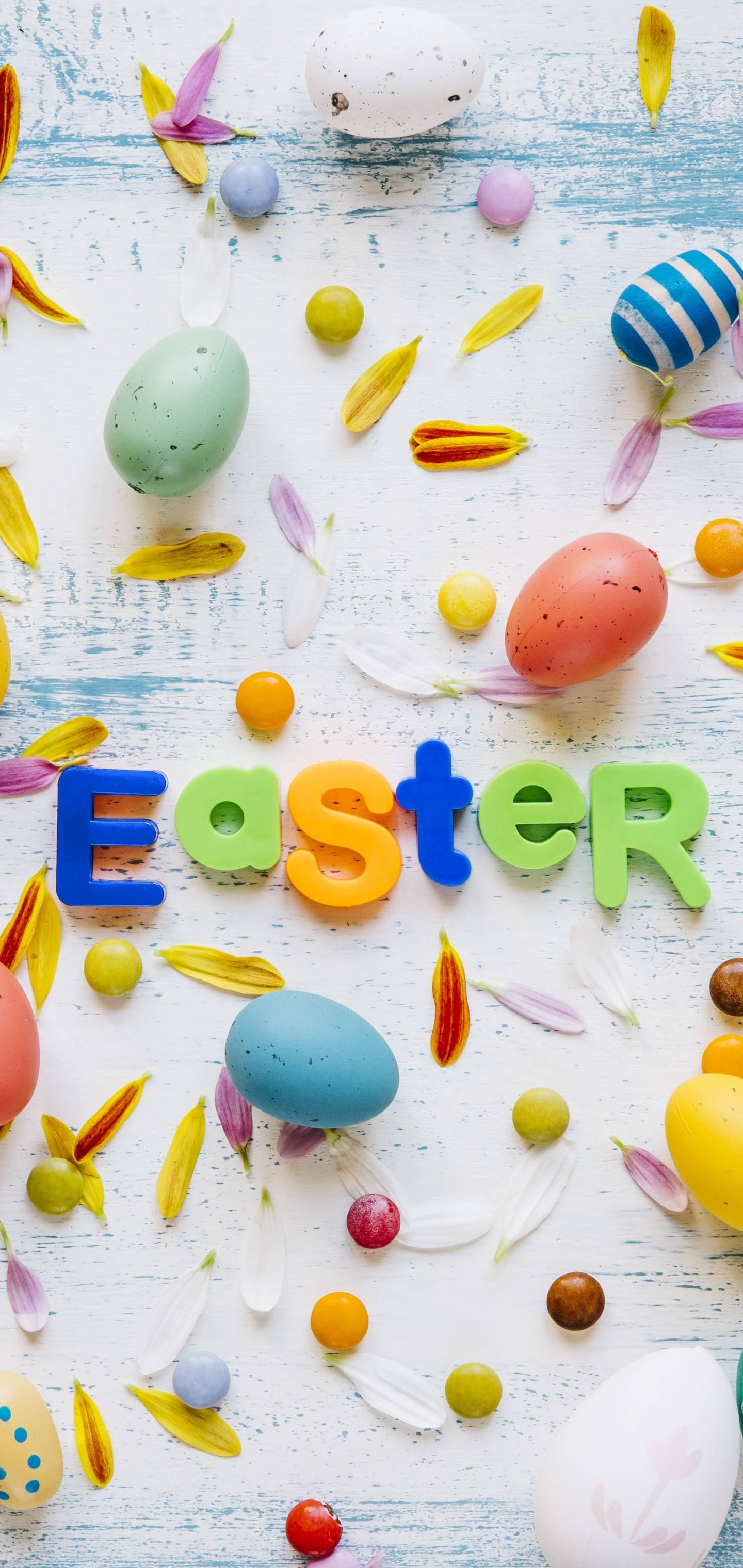 Easter Phone 桌布 壁紙 — 色彩繽紛的復活節蛋和花瓣散落在風化的藍色木質表面上，並用明亮的字母拼出Easter一詞。