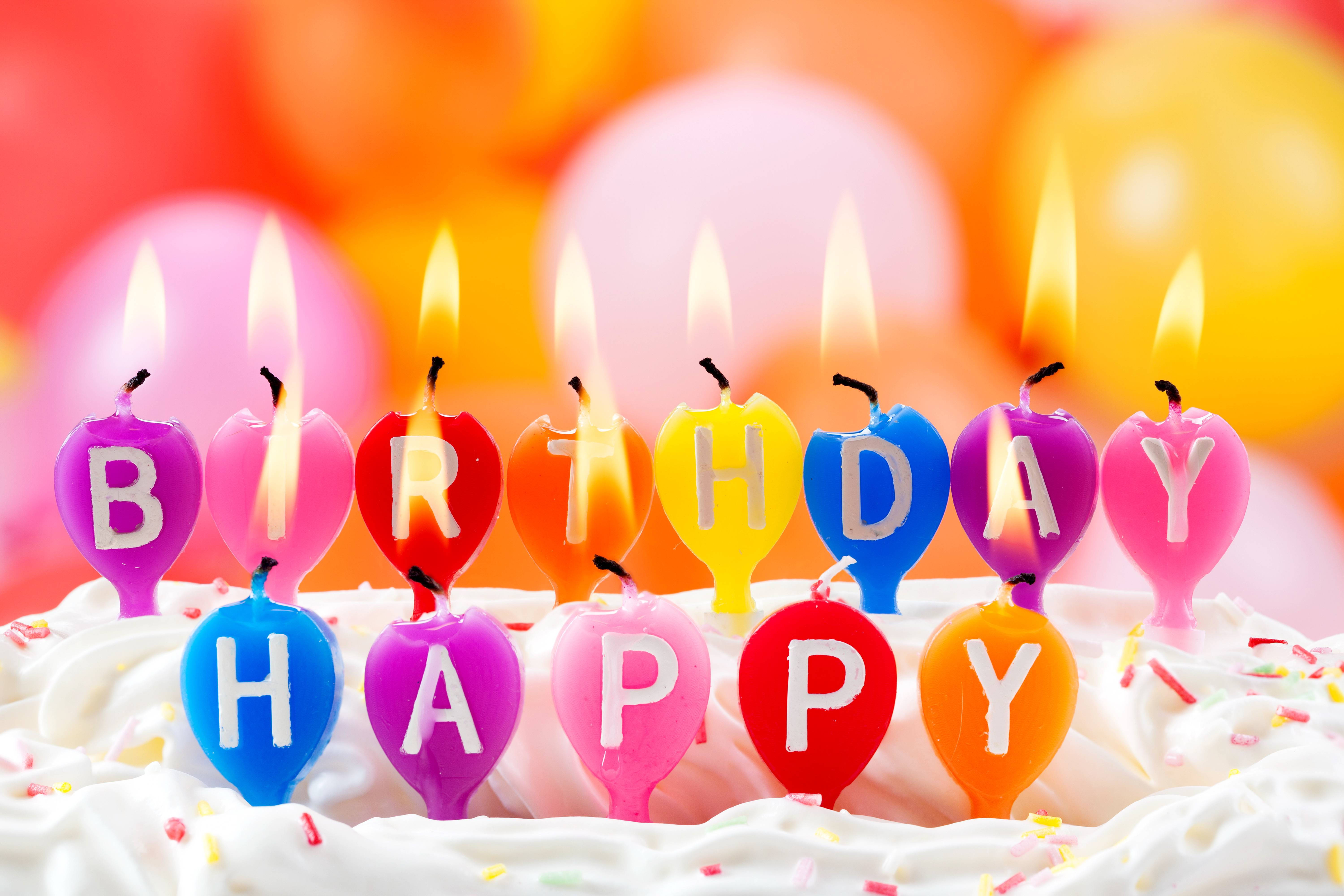 生日快樂 桌布 壁紙 — 色彩繽紛的心形生日蠟燭拼出「HAPPY BIRTHDAY」，在糖霜蛋糕上閃耀，蛋糕上有彩色糖粒，背景有模糊的氣球。