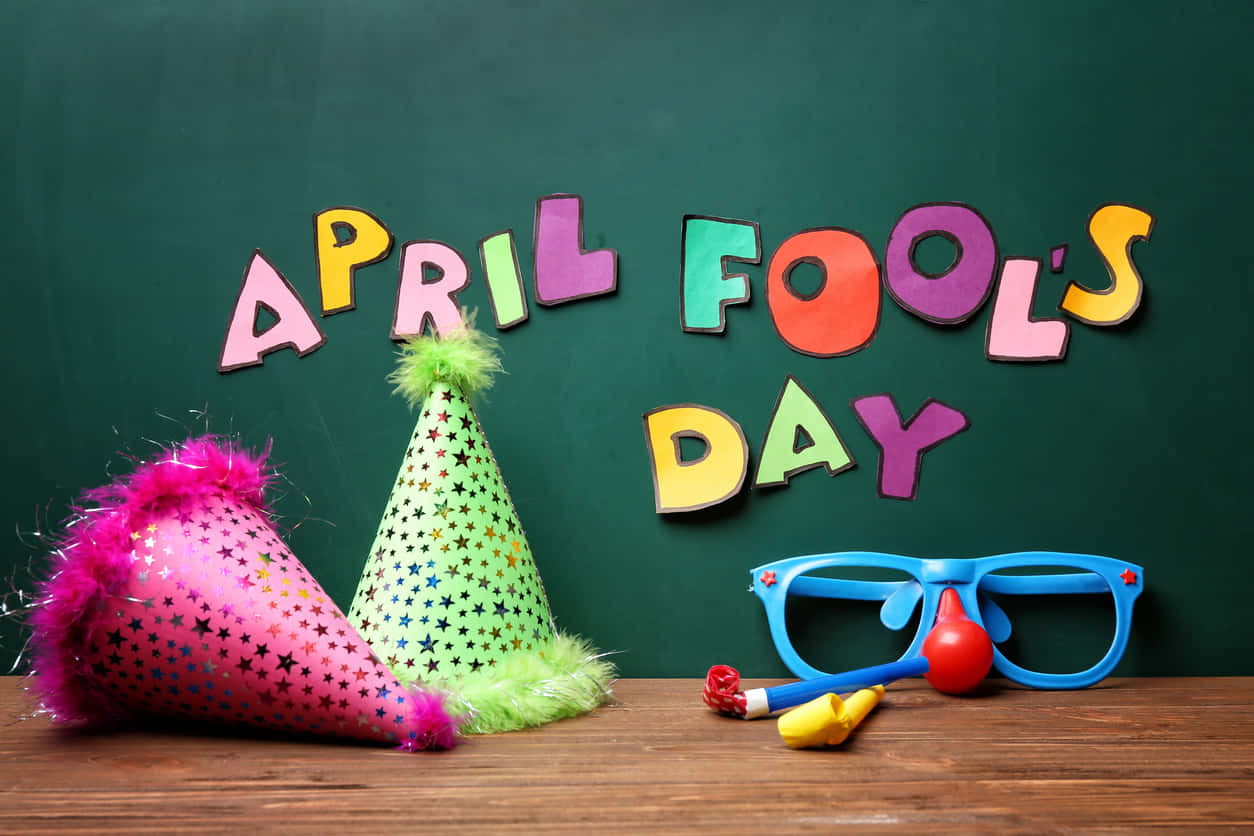 April Fools 桌布 壁紙 — 色彩繽紛的愚人節字母和派對道具如帽子和眼鏡，放在木桌上，背景是綠色黑板。