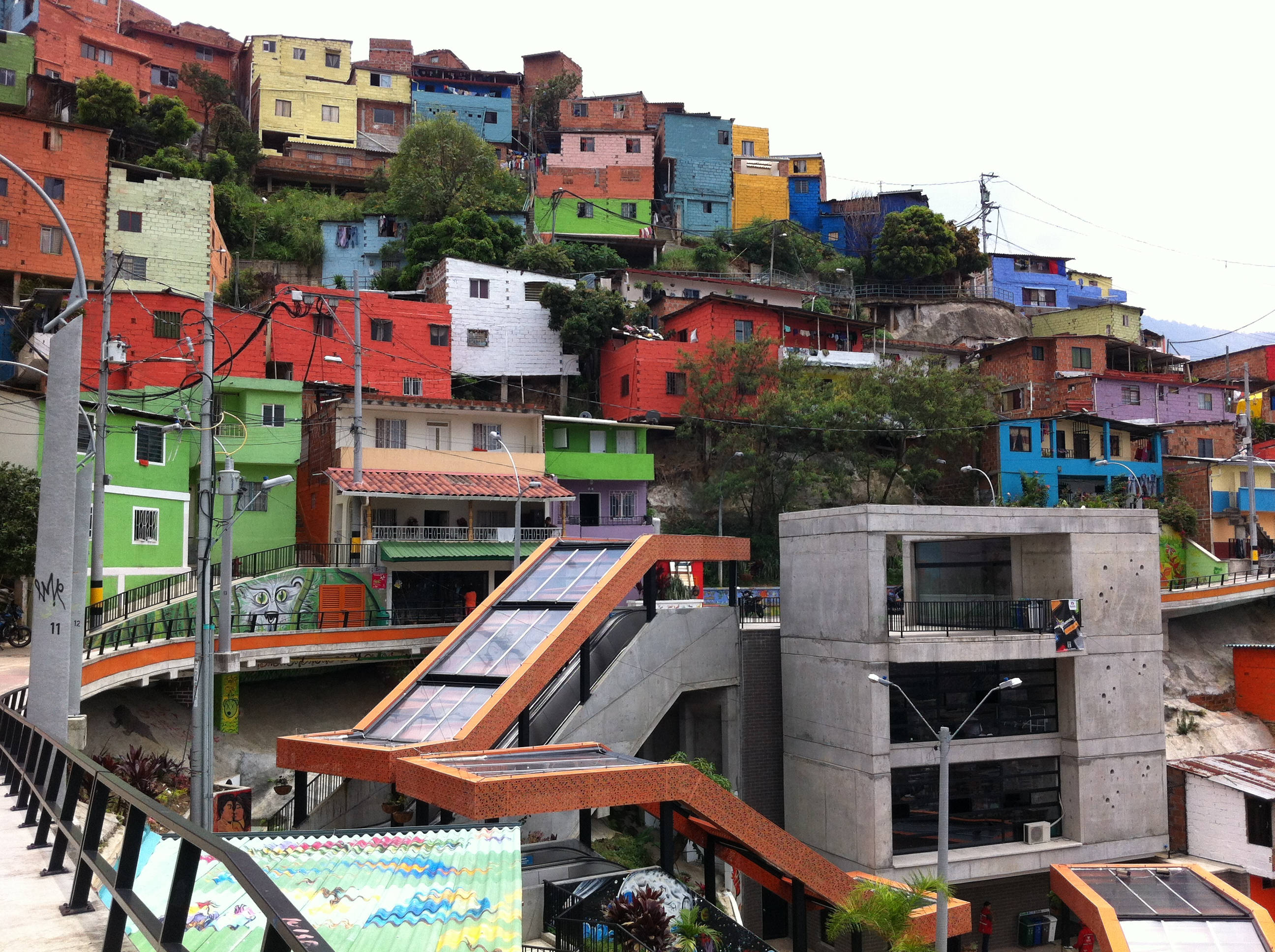 Medellin 桌布 壁紙 — 色彩繽紛的房屋沿著陡峭的山坡層疊而下，旁邊是現代混凝土建築和自動扶梯。